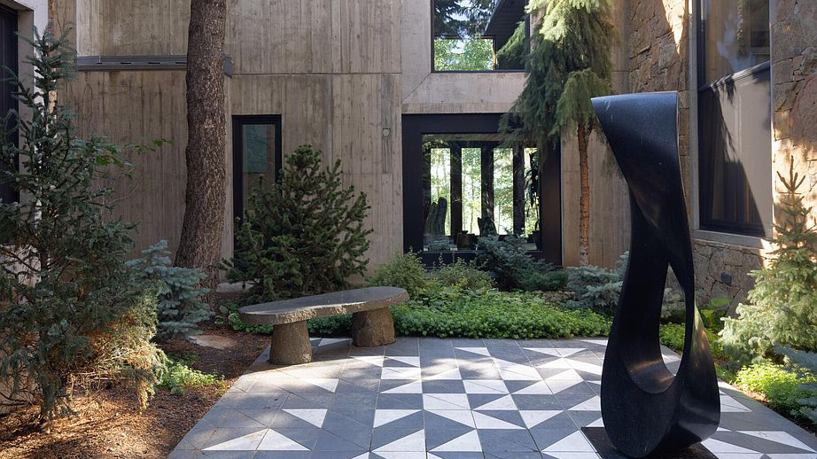 This image depicts a well-manicured garden area adjacent to a modern home. A striking black sculpture stands on a geometric tiled patio, complemented by a stone bench surrounded by lush greenery and small trees. The architectural design is modern, with concrete and stone elements, suggesting a serene and sophisticated outdoor space perfect for relaxation or entertaining.
