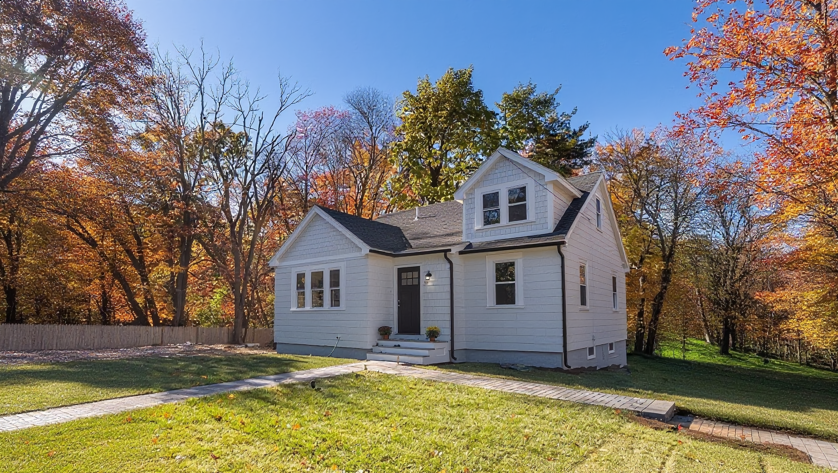 This is a charming front exterior view of a single-family home. The house is painted in a light color, accented by a dark front door and black gutters. The yard is well-manicured with a stone walkway leading to the entrance, and colorful autumn foliage in the background enhances the curb appeal.