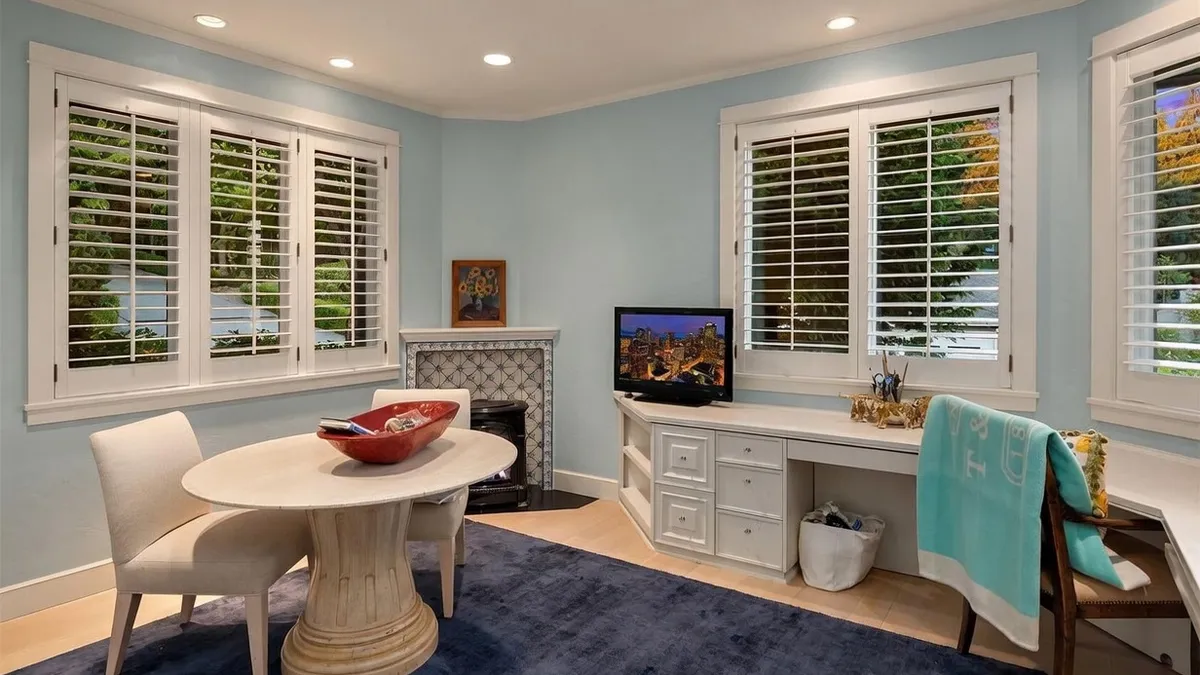 This light-filled multi-purpose room features a functional corner workspace with built-in cabinetry and a television, alongside an adjacent round dining or casual table. The space is accented with soft blue walls, plantations shutters, and a decorative fireplace niche, creating a versatile and inviting atmosphere. A plush dark blue rug grounds the room, blending office productivity with comfortable living elements.