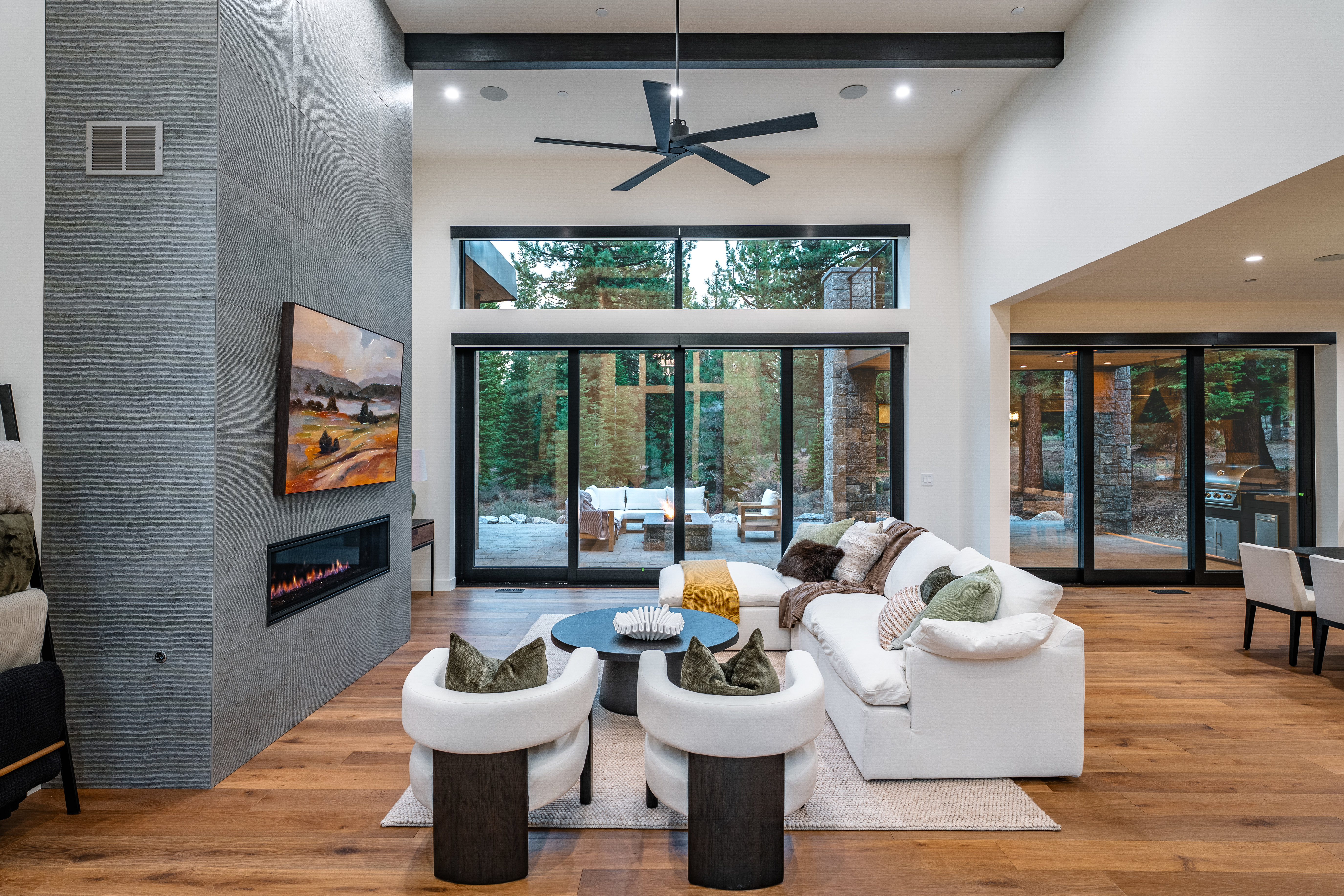This is a bright and airy living room with a modern design. The room features a floor-to-ceiling stone fireplace, a large sectional sofa, and floor-to-ceiling windows that offer views of the surrounding trees. The hardwood floors and neutral color palette create a warm and inviting atmosphere.