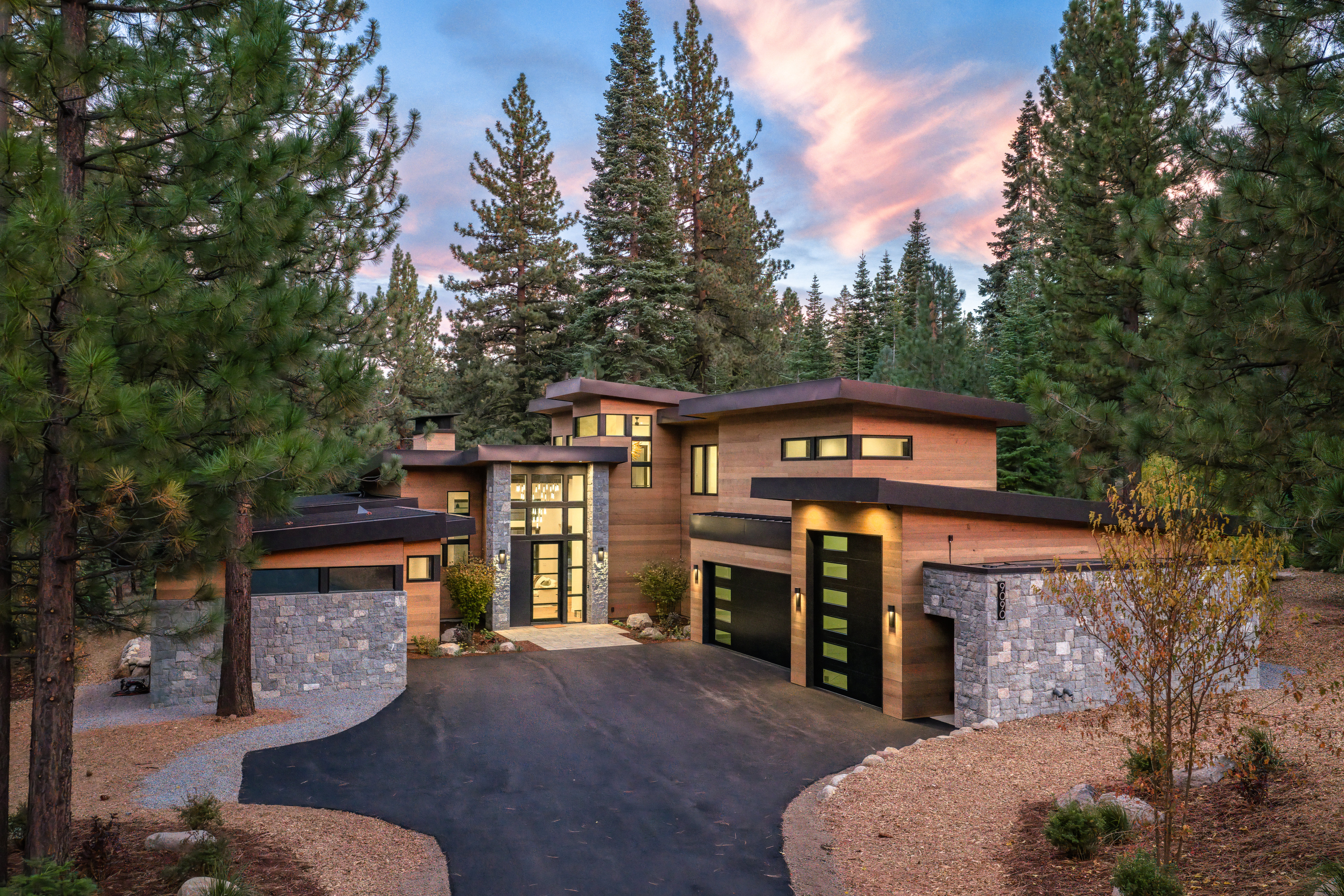 This is a striking front view of a modern home, showcasing its unique architectural design with a combination of wood and stone elements. The property features a well-manicured driveway leading to a multi-car garage and a grand entrance with large windows. The surrounding mature trees and the colorful sky at dusk create a serene and luxurious atmosphere.