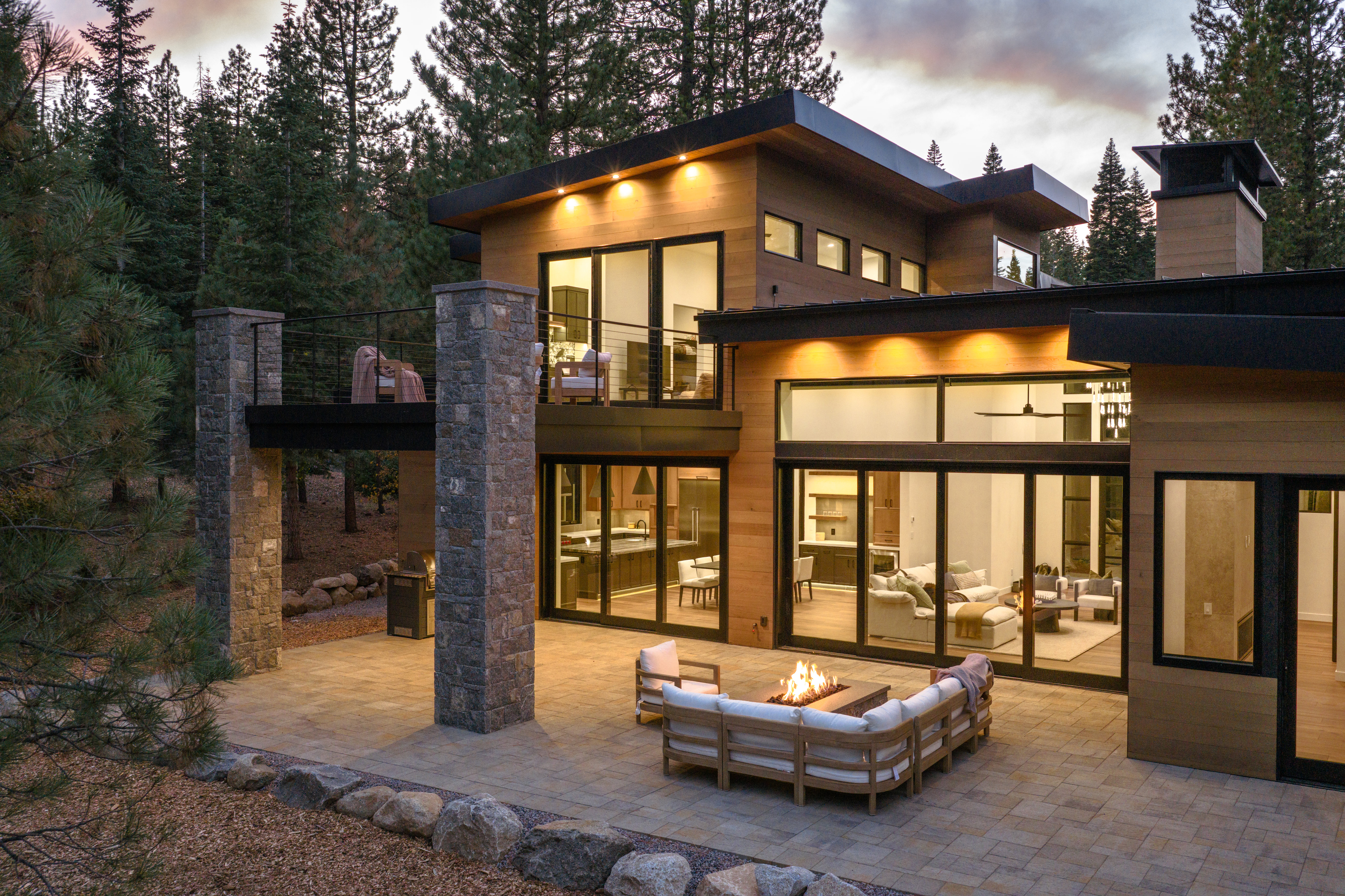 This is a rear exterior view of a modern home, showcasing a spacious patio with a fire pit and comfortable seating. Large glass doors provide seamless indoor-outdoor living, revealing glimpses of the interior living spaces and kitchen. The architectural design features clean lines, wood siding, and stone accents, blending harmoniously with the surrounding forest landscape.