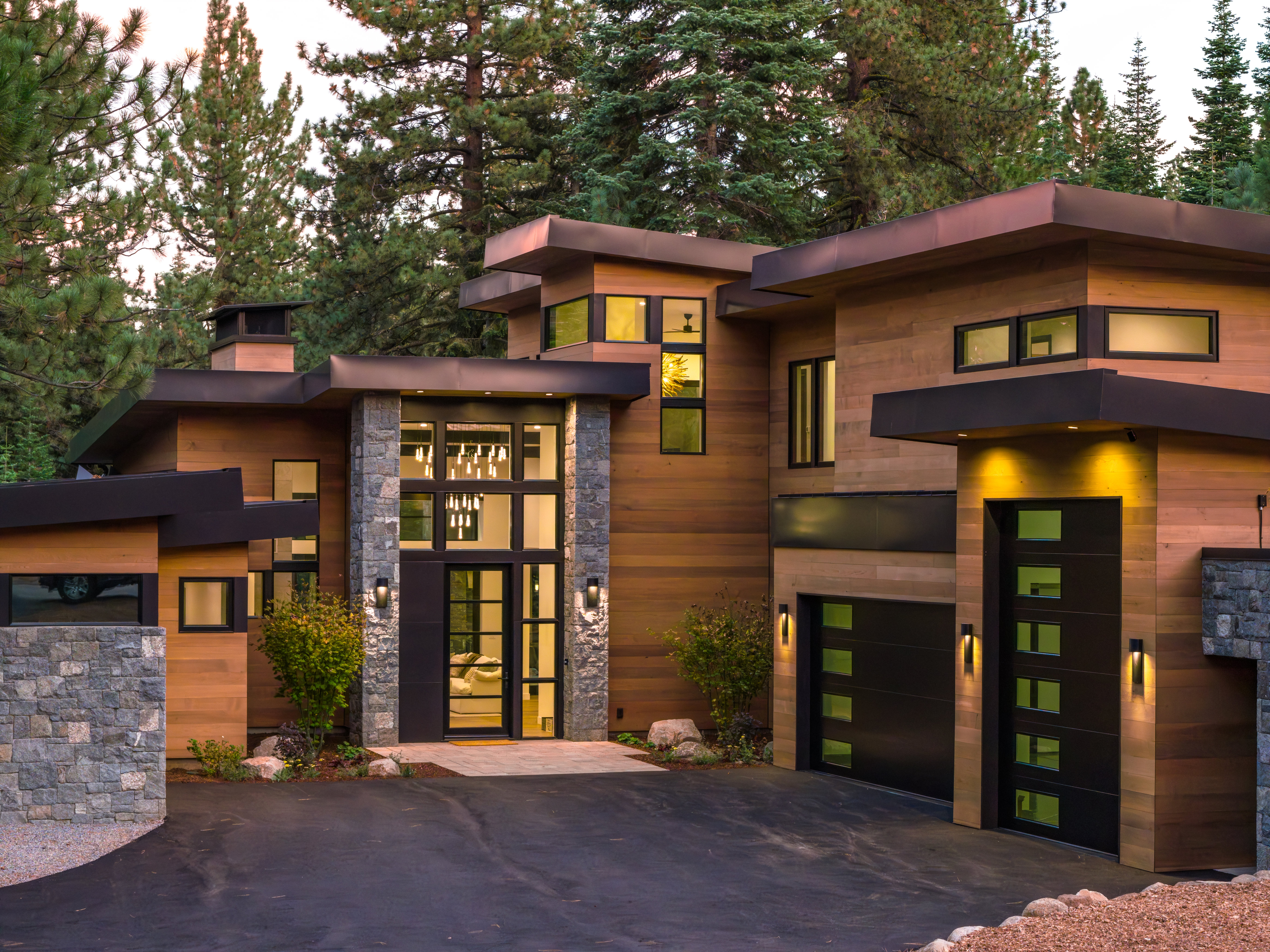 This is a front view of a modern, luxurious home with a wood and stone facade. The house features a flat roof, black-framed windows, and a prominent entryway with a glass door and sidelights. A paved driveway leads to the two-car garage, and the landscaping includes trees and shrubs, creating a sense of privacy and elegance.