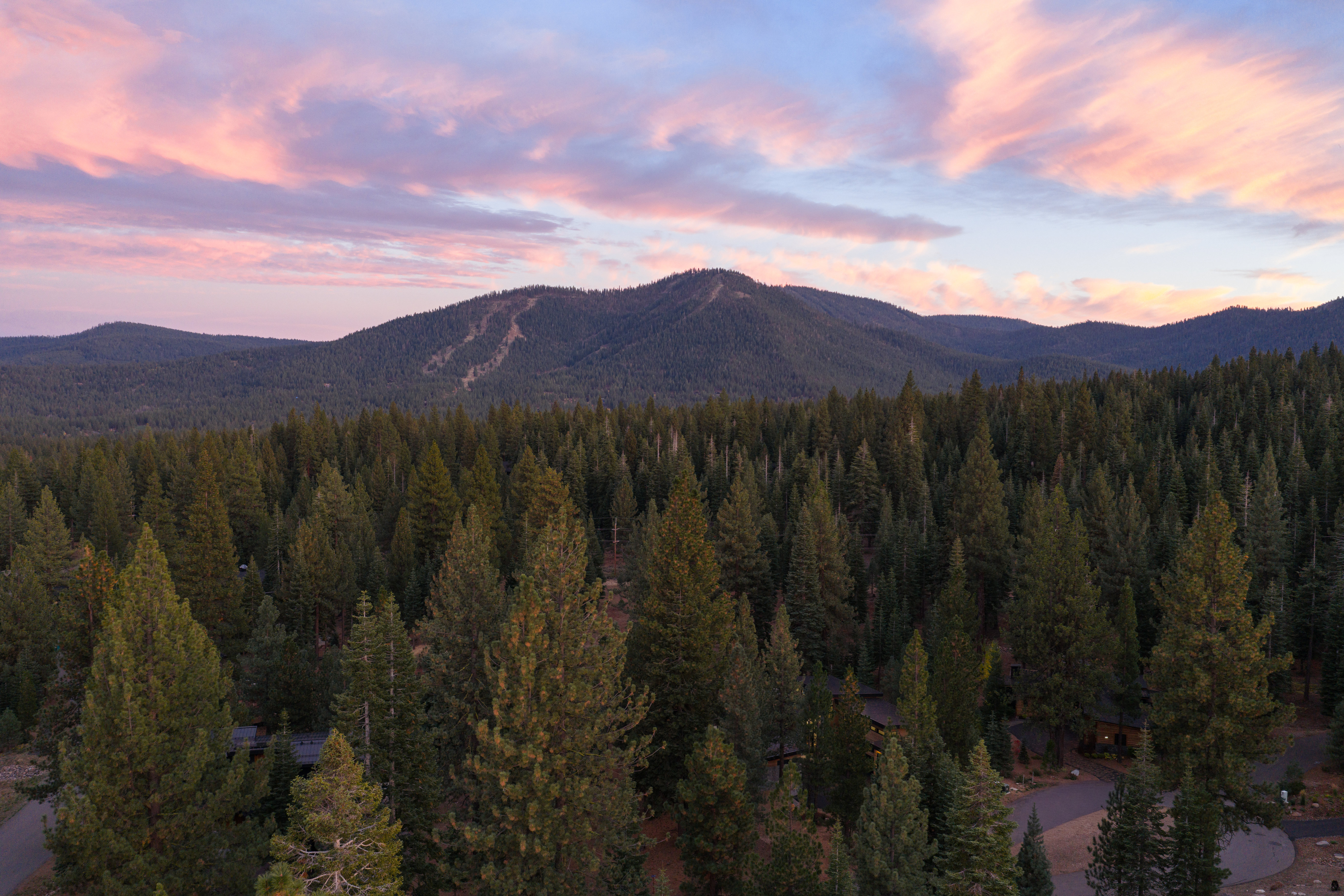 This aerial view showcases a property nestled within a dense forest, with a mountain range visible in the background under a sky painted with hues of pink and blue at dusk. The landscape emphasizes privacy and natural beauty, suggesting a secluded and serene living environment. The presence of mature trees and the distant mountain view add to the property's appeal, highlighting its connection to nature.