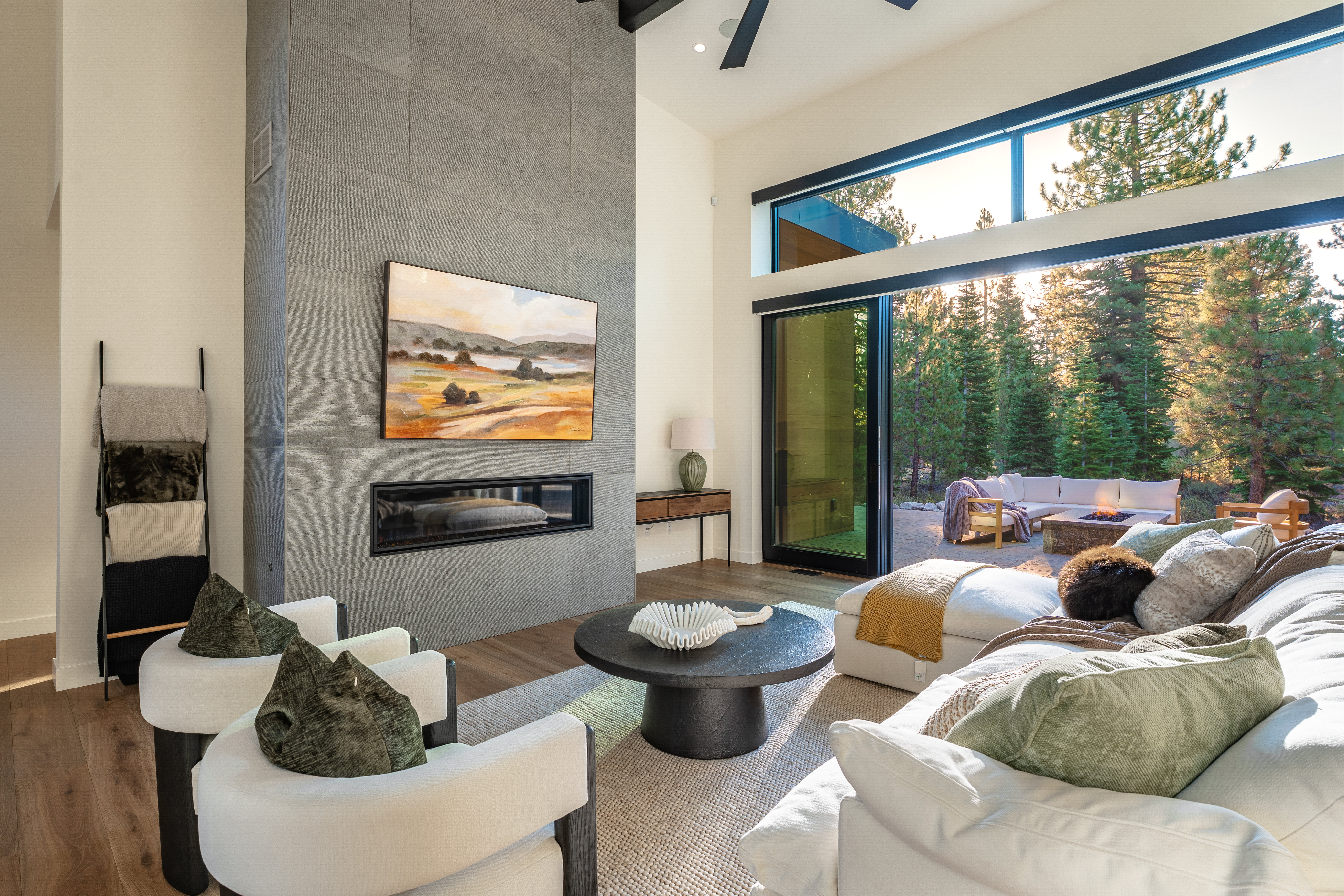 This is a bright and airy living room featuring a modern fireplace with a gray stone surround and a large painting above. The room is furnished with comfortable white sofas and chairs, a round coffee table, and a ladder with blankets. Sliding glass doors open to an outdoor patio with additional seating and a fire pit, seamlessly blending indoor and outdoor living.