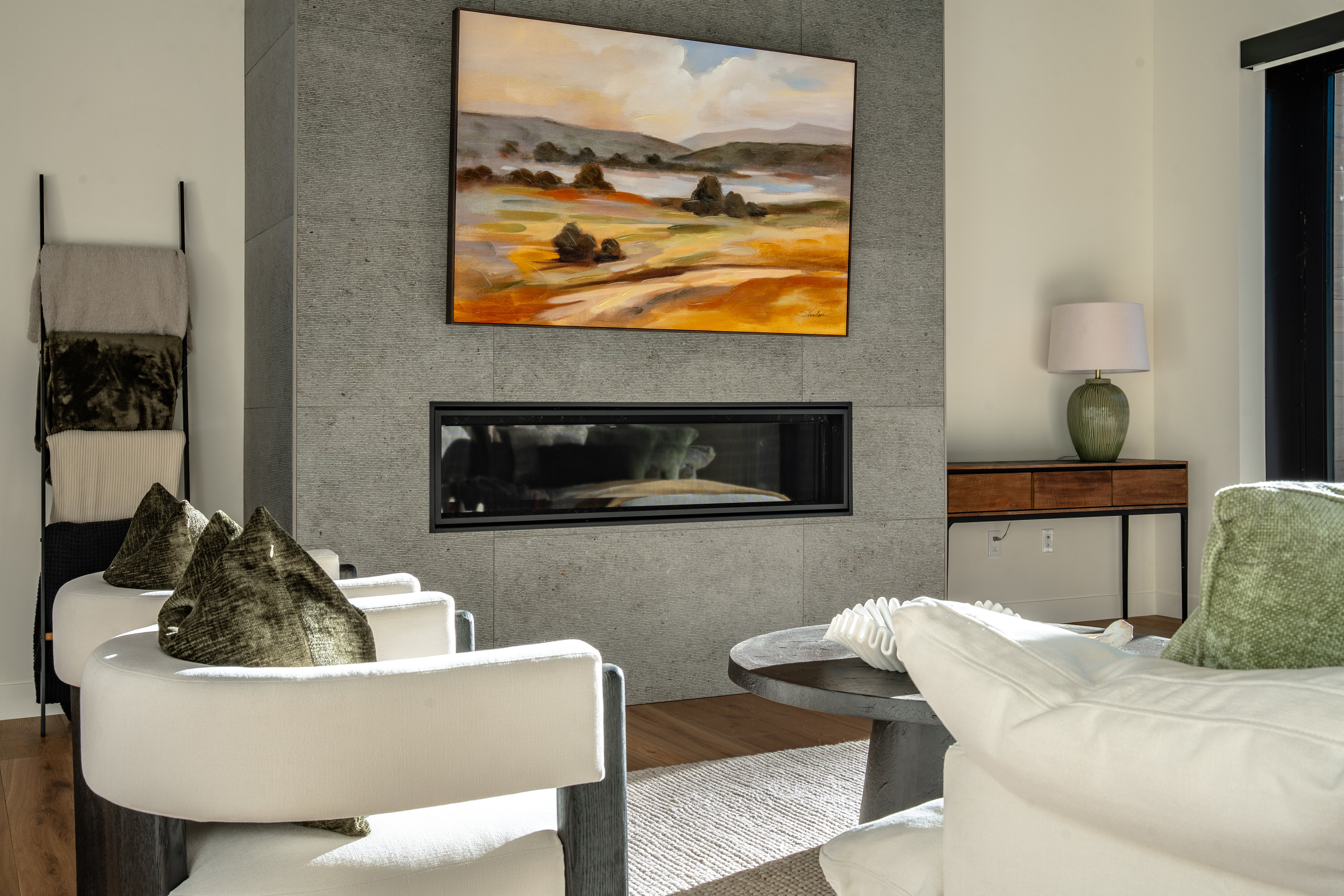This living room showcases a modern fireplace with a gray stone surround and a contemporary art piece above. Two white armchairs with dark wood frames face the fireplace, complemented by a dark wood coffee table and a light-colored rug. The room exudes a sophisticated and comfortable ambiance, perfect for relaxation and entertaining.