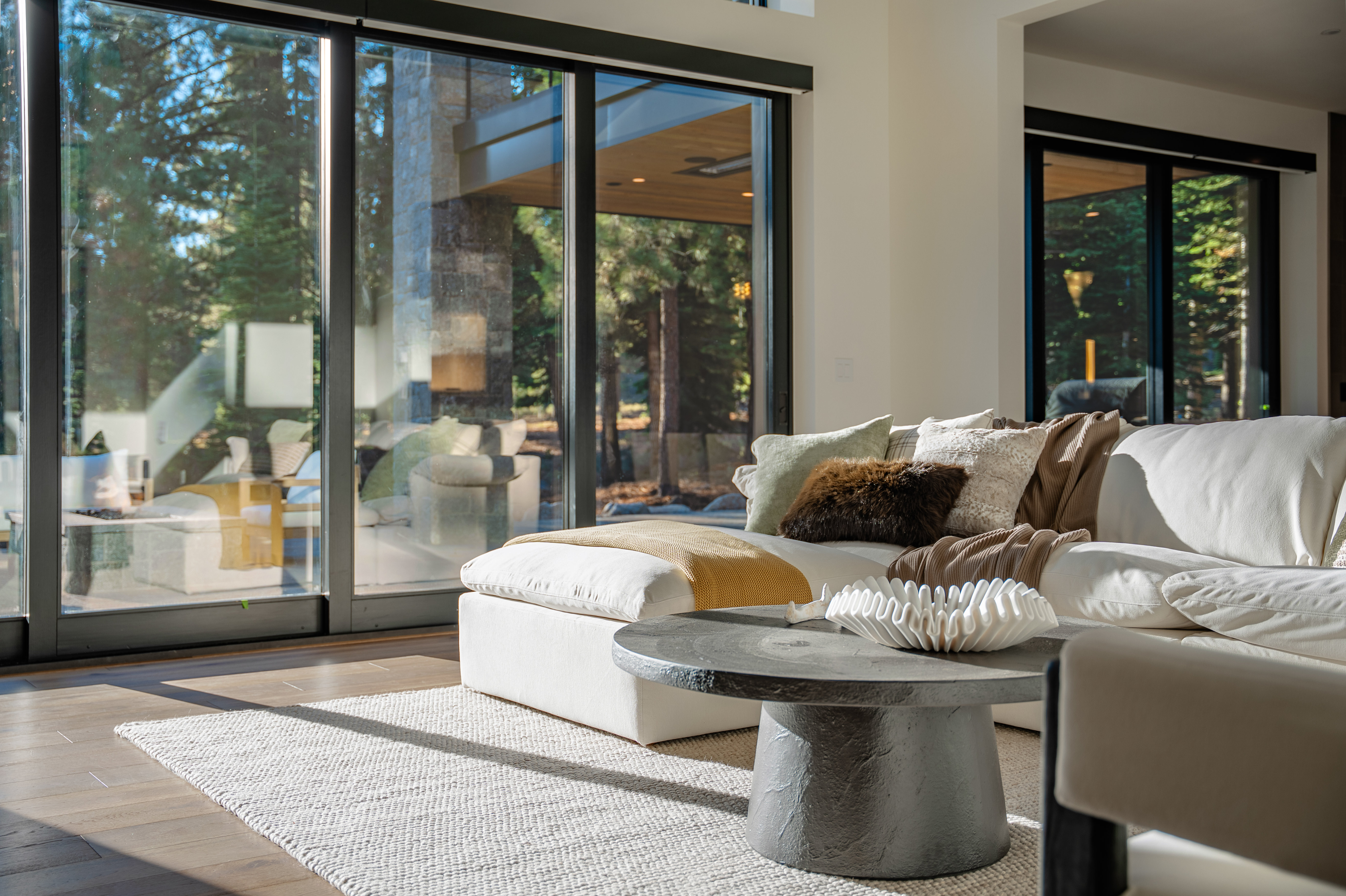 This is a bright and airy living room featuring a large sectional sofa adorned with decorative pillows and a throw blanket. A modern coffee table with a sculptural centerpiece sits on a light-colored area rug. Large sliding glass doors offer views of the surrounding trees and bring natural light into the space, creating a seamless indoor-outdoor living experience.