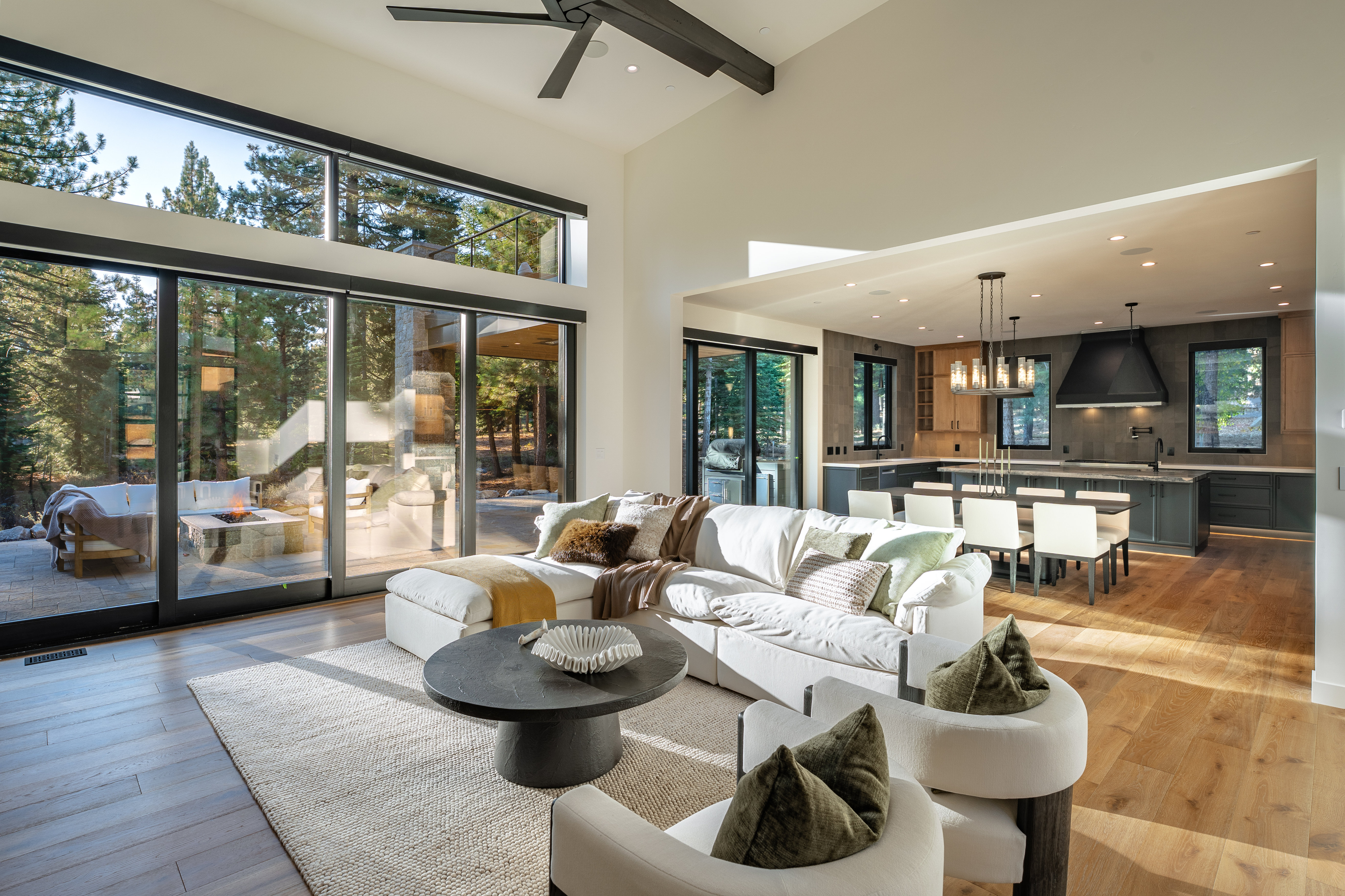 This is a bright and airy living room featuring a large sectional sofa, modern armchairs, and a round coffee table. Expansive windows offer views of the surrounding trees and outdoor patio area. The open floor plan seamlessly connects the living room to the kitchen and dining area, creating a spacious and inviting atmosphere.