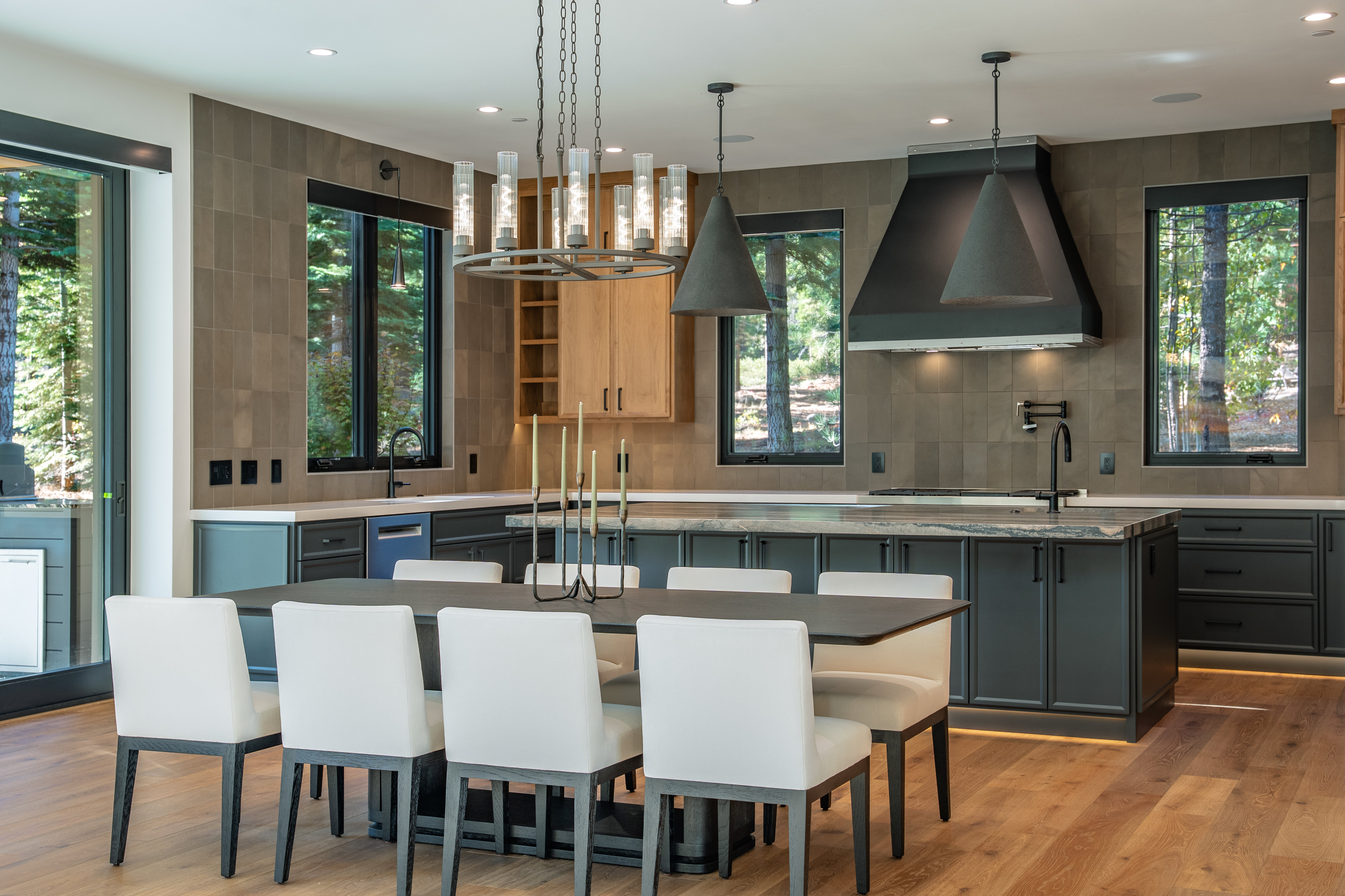 This is a modern kitchen with dark cabinetry, a large island with a unique countertop, and stainless steel appliances. The kitchen features a dining table with white chairs, a statement chandelier, and pendant lighting above the island. Large windows offer views of the surrounding trees, creating a connection with nature.