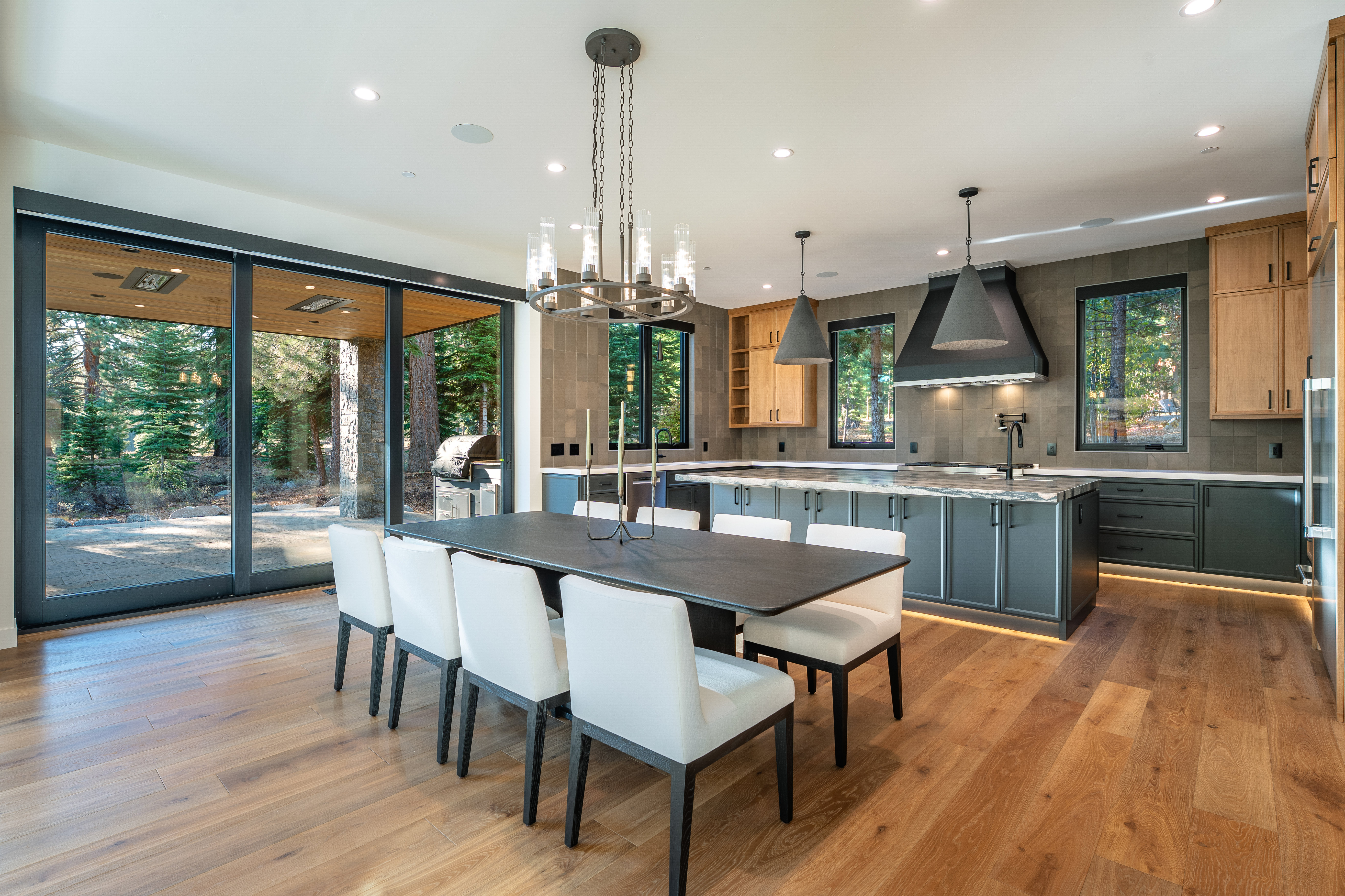 This is a well-lit, modern kitchen and dining area featuring a dark wood dining table with white upholstered chairs, a contemporary chandelier, and a kitchen island with dark cabinetry and light countertops. The kitchen includes stainless steel appliances, wood cabinets, and a dark range hood, with a large sliding glass door providing natural light and a view of the outdoor area. The hardwood floors add warmth to the space, creating an inviting and luxurious atmosphere.