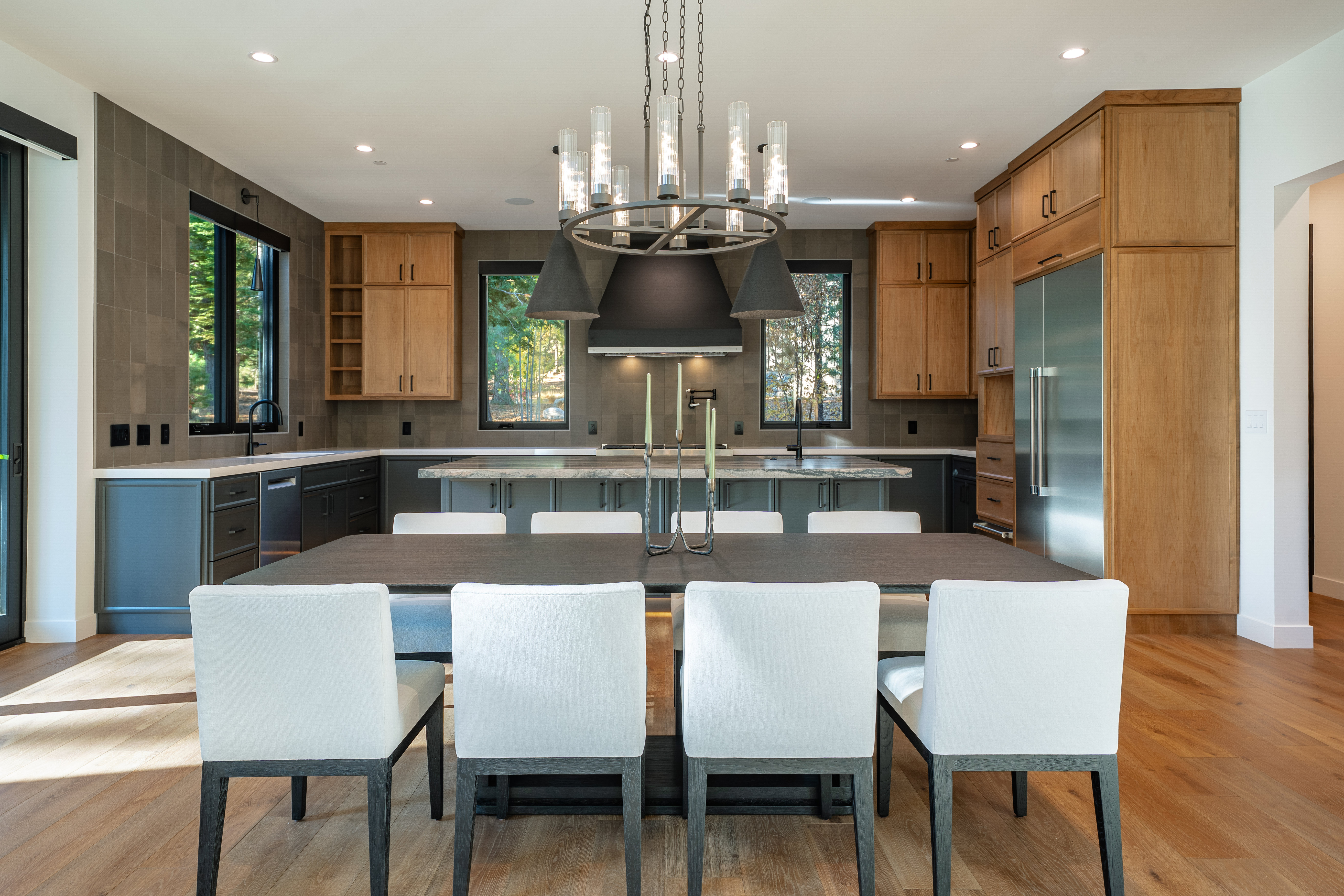 This is a well-lit, modern kitchen featuring a large dining table with white chairs in the foreground. The kitchen includes dark lower cabinets, light wood upper cabinets, stainless steel appliances, and a central island with a light countertop. A statement chandelier hangs above the dining table, and windows provide natural light and views to the outside.