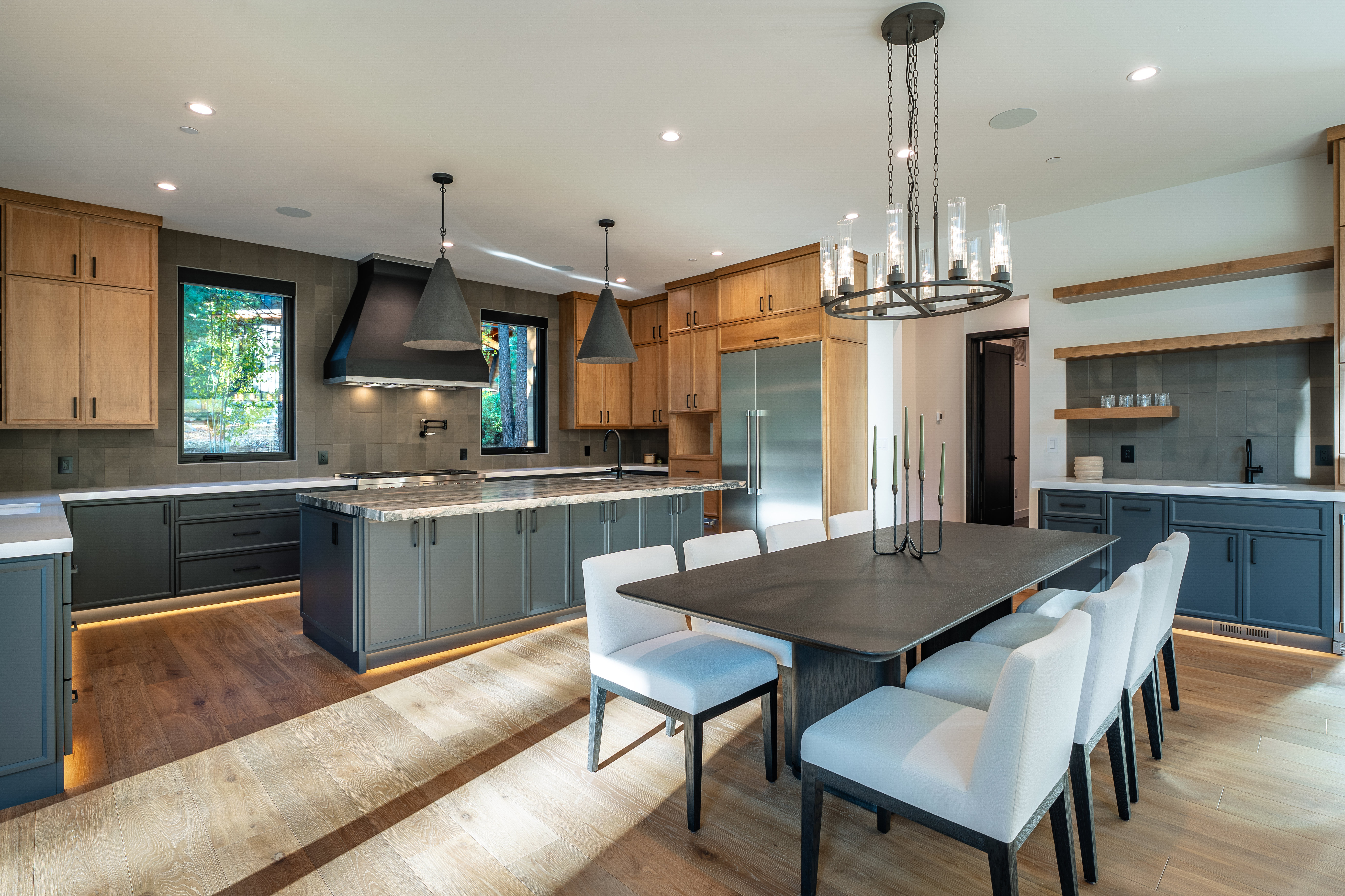 This is a spacious, modern kitchen and dining area featuring a large island with a marble countertop, custom cabinetry in two tones (wood and gray), and stainless steel appliances. A dining table with white chairs sits adjacent to the kitchen island, illuminated by a modern chandelier. The room is well-lit with recessed lighting and natural light from the windows, creating a warm and inviting atmosphere.