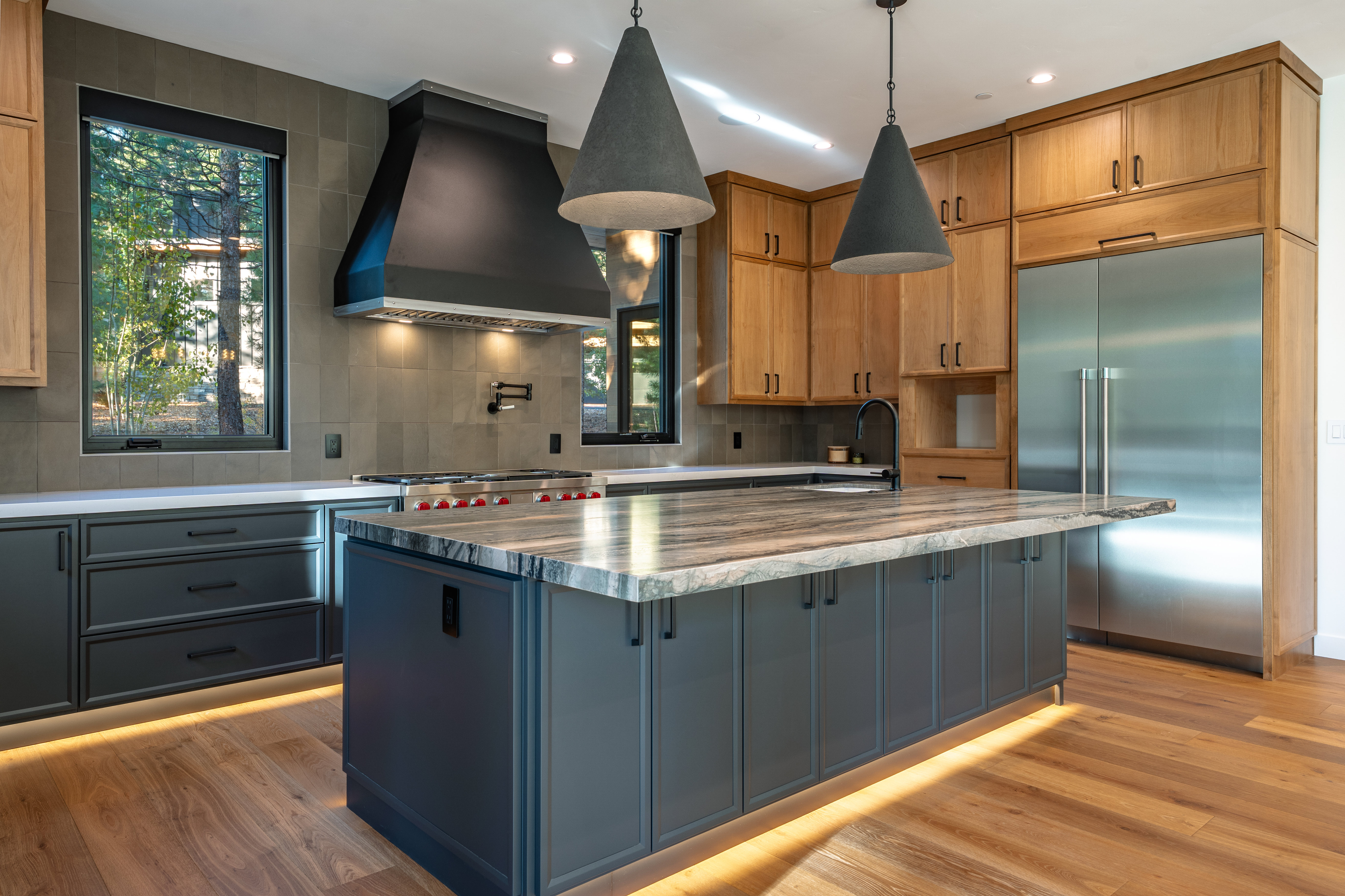 This is a well-lit, modern kitchen featuring a large island with a marble countertop and dark gray cabinetry. The kitchen also includes stainless steel appliances, light wood cabinets, and a black range hood. The hardwood flooring adds warmth to the space, and pendant lights hang above the island.
