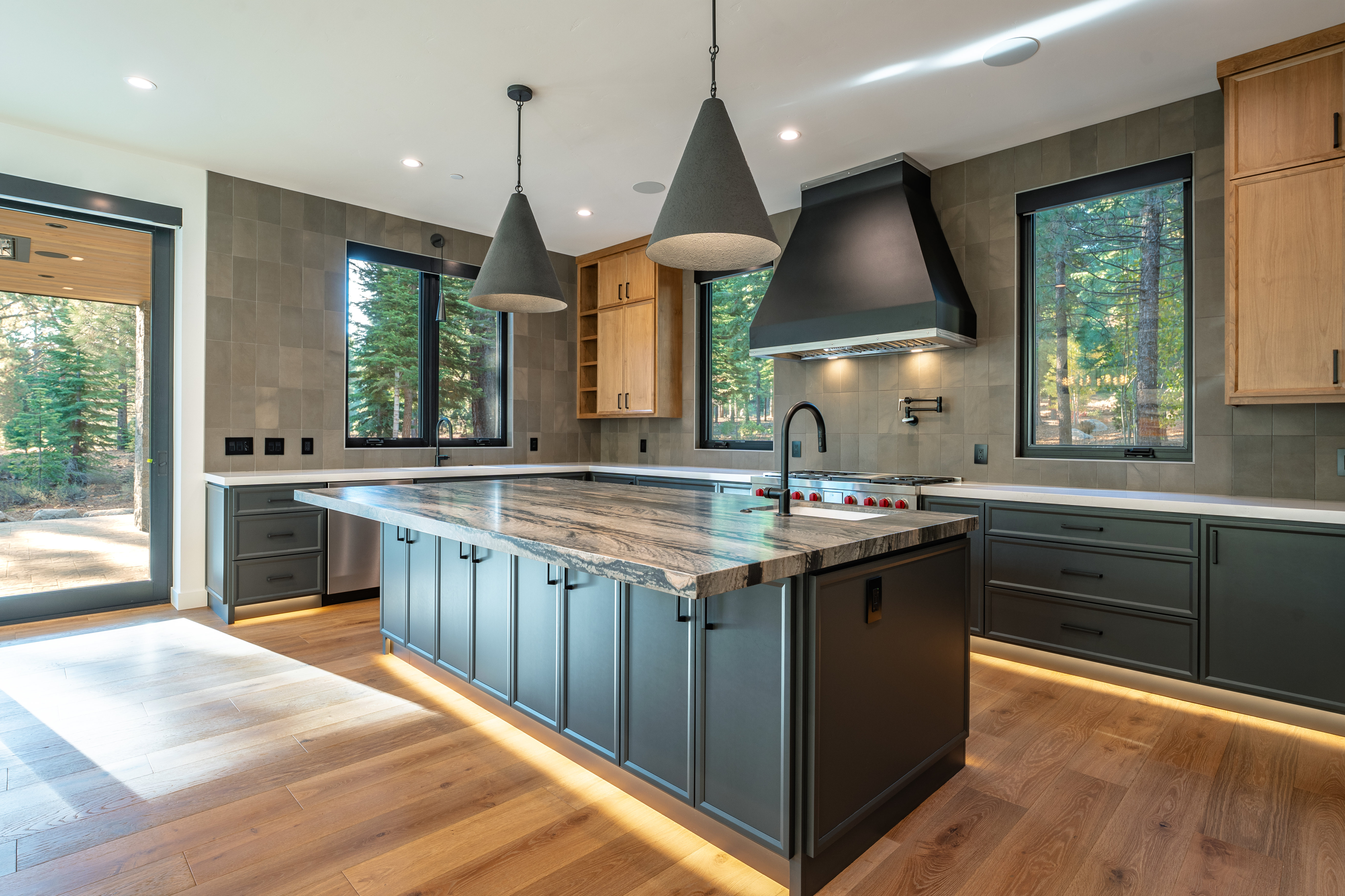 This is a well-lit, modern kitchen featuring a large island with a unique granite countertop and dark gray cabinetry. Stainless steel appliances are integrated seamlessly, and pendant lights hang above the island. Large windows offer views of the surrounding trees, creating a connection with nature.