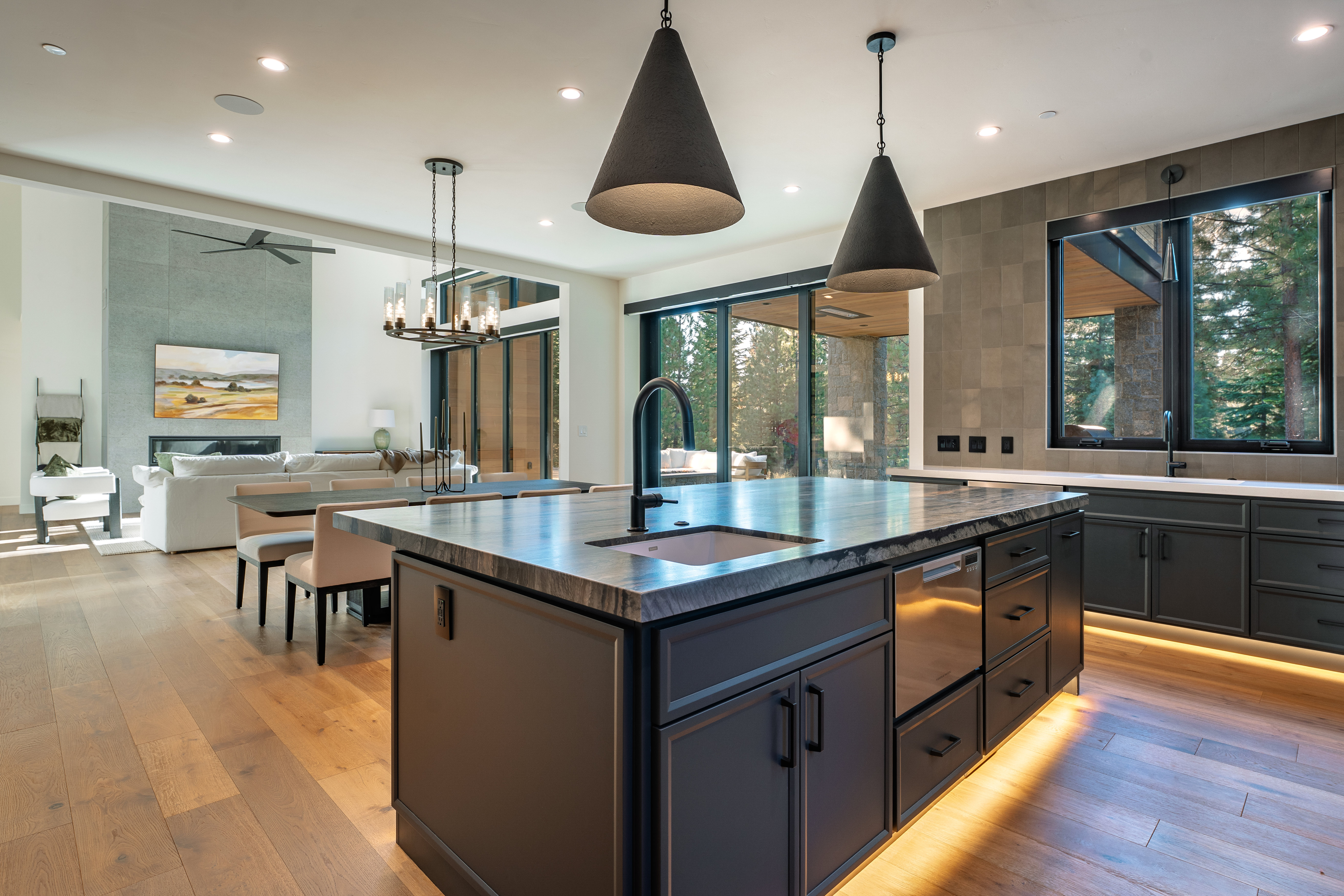 This is a modern, open-concept kitchen featuring a large island with a dark countertop and sink, complemented by dark cabinetry and stainless steel appliances. Pendant lights hang above the island, and large windows offer views of the outdoors, creating a bright and inviting space that seamlessly connects to the dining and living areas. Under-cabinet lighting adds a warm glow, highlighting the hardwood floors.
