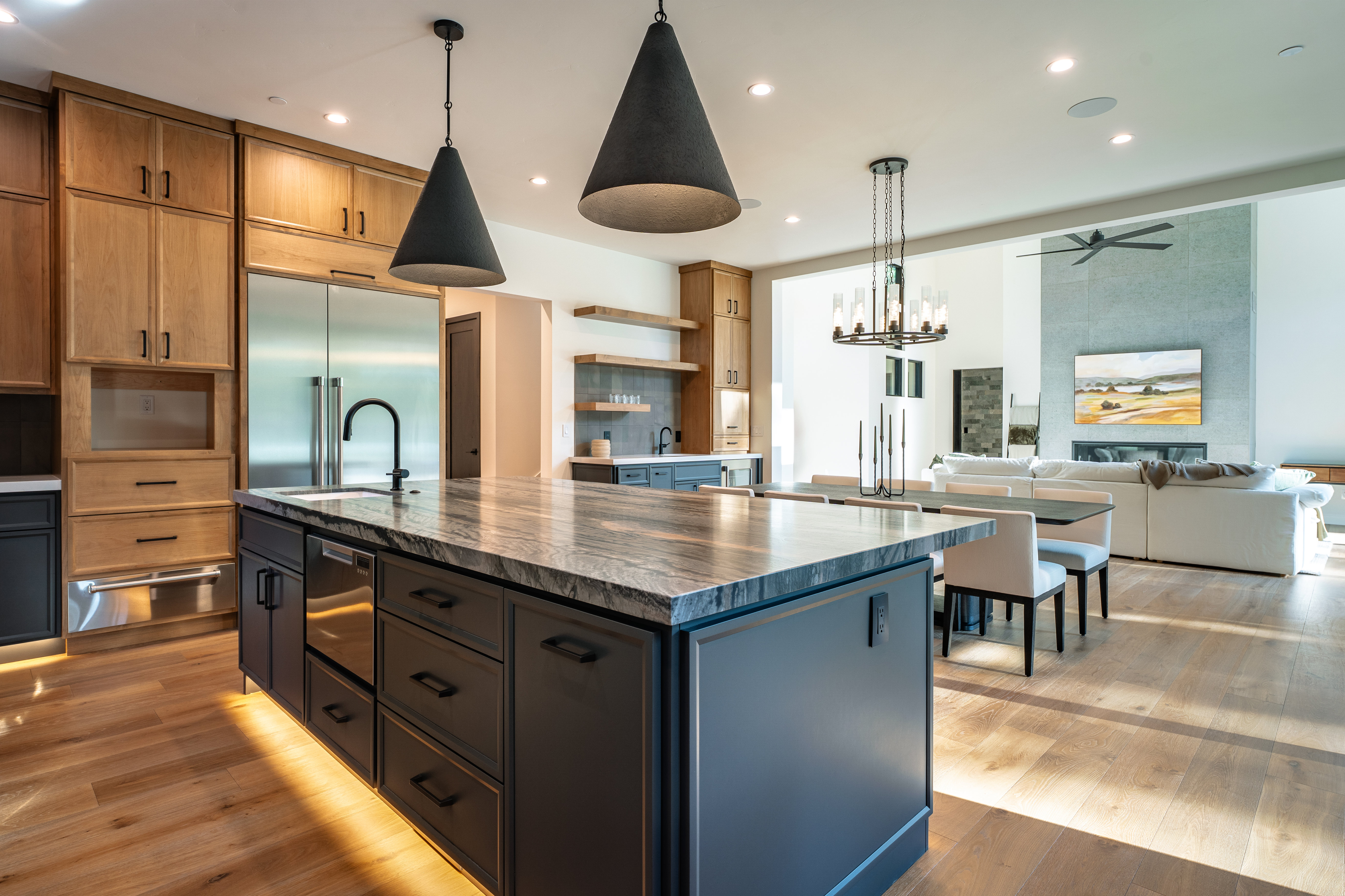 This is a spacious, modern kitchen featuring a large island with a marble countertop and dark cabinetry. The kitchen also includes light wood cabinets, stainless steel appliances, and unique pendant lighting. The open floor plan seamlessly connects the kitchen to the dining and living areas, creating an inviting and luxurious atmosphere.