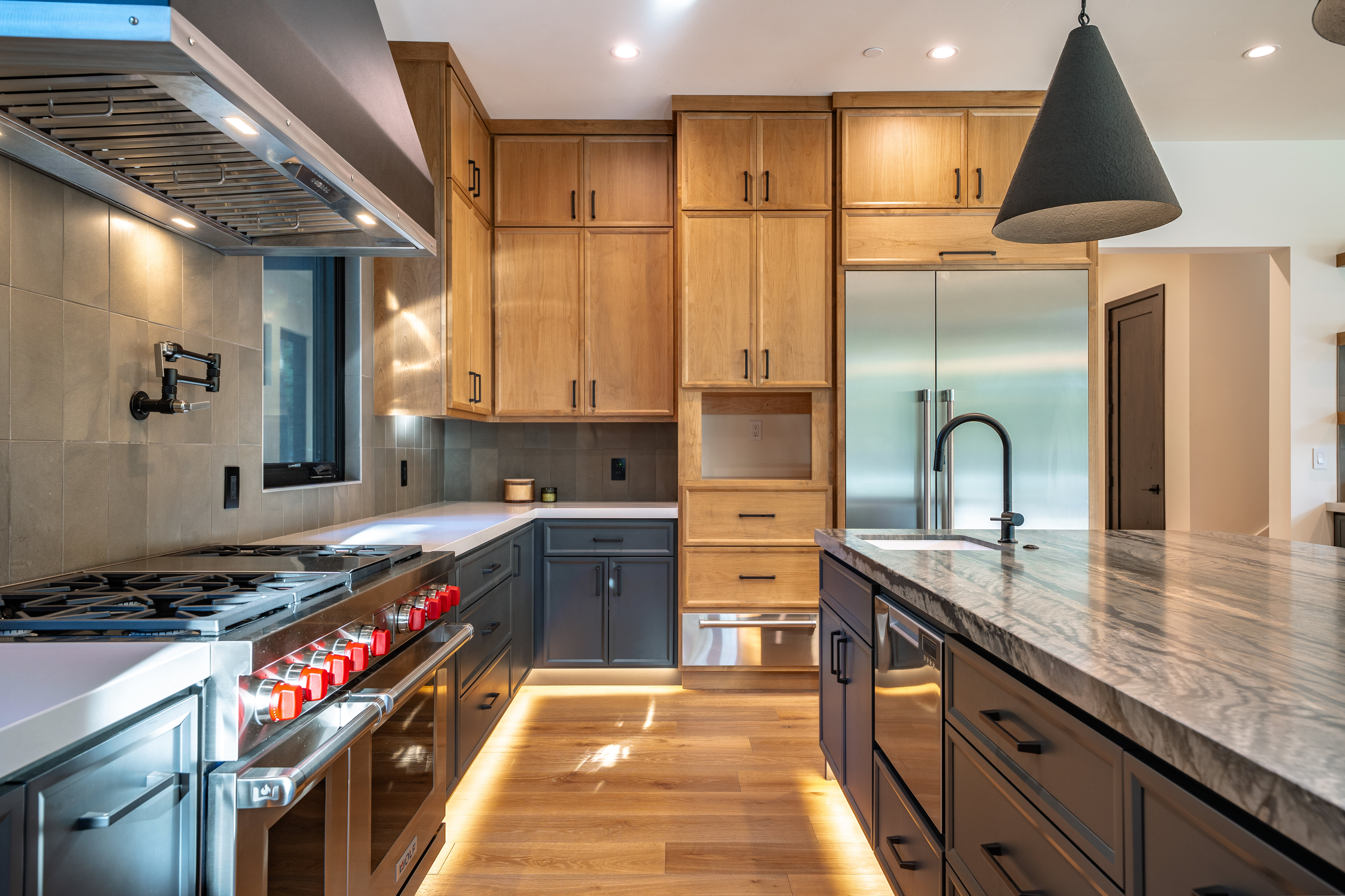 This is a well-lit, modern kitchen featuring a stainless steel range with red knobs and a stainless steel hood. The kitchen has a mix of light wood and dark gray cabinetry, with a large island featuring a marble countertop and a black faucet. Under-cabinet lighting and a dark pendant light add to the ambiance.