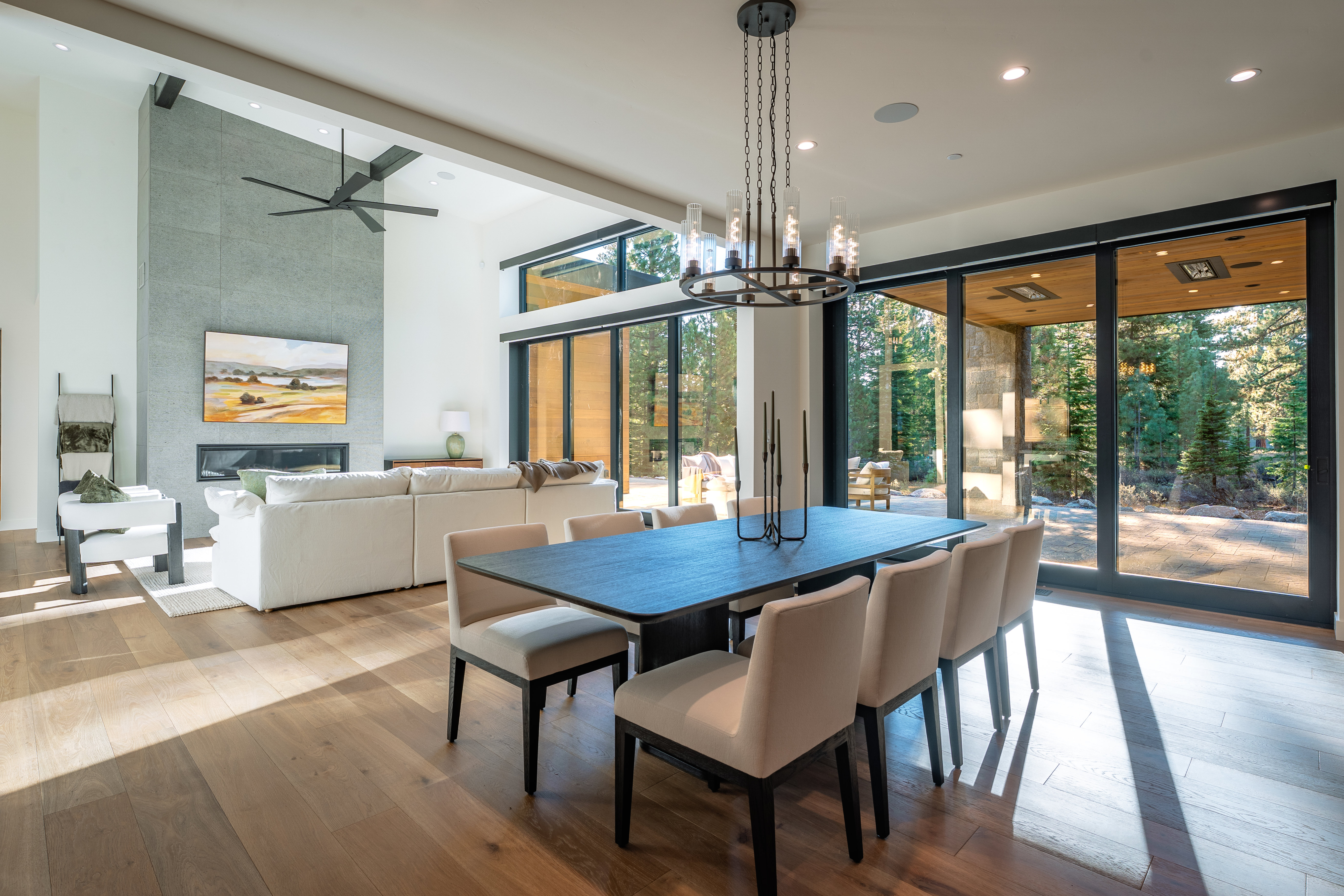 This is an interior shot of a modern dining room. The room features a long, dark wood dining table surrounded by upholstered chairs with dark wood legs. A contemporary chandelier hangs above the table, and large sliding glass doors provide ample natural light and a view of the outdoor scenery. The flooring is light wood, and the overall aesthetic is clean, sophisticated, and inviting.