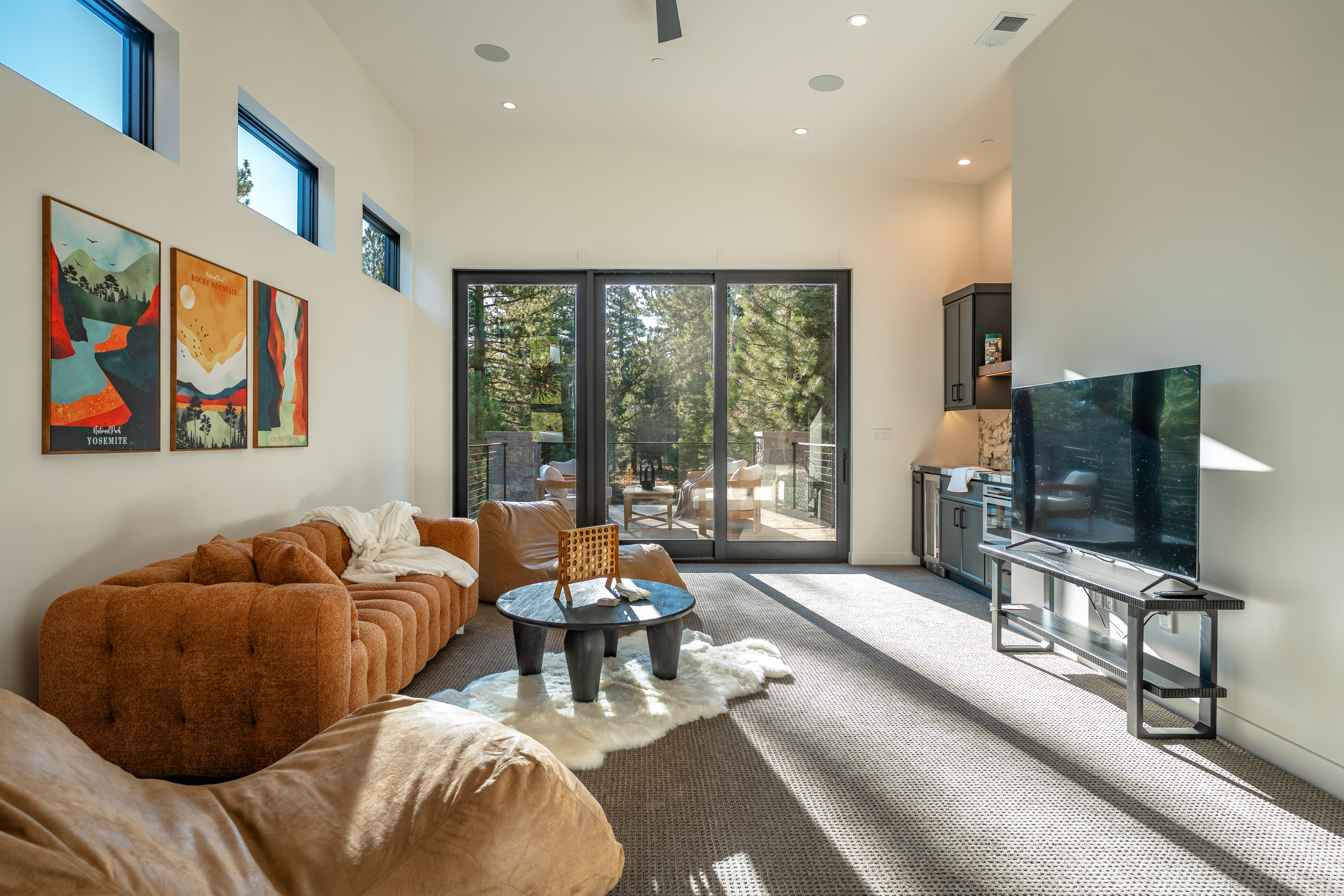 This is a bright and modern living room featuring a comfortable brown sofa, a unique round coffee table on a white sheepskin rug, and a large sliding glass door leading to an outdoor deck. The room is decorated with artwork and includes a built-in bar area with a television, creating a cozy yet sophisticated atmosphere. The perspective is from a low angle, emphasizing the depth and spaciousness of the room.