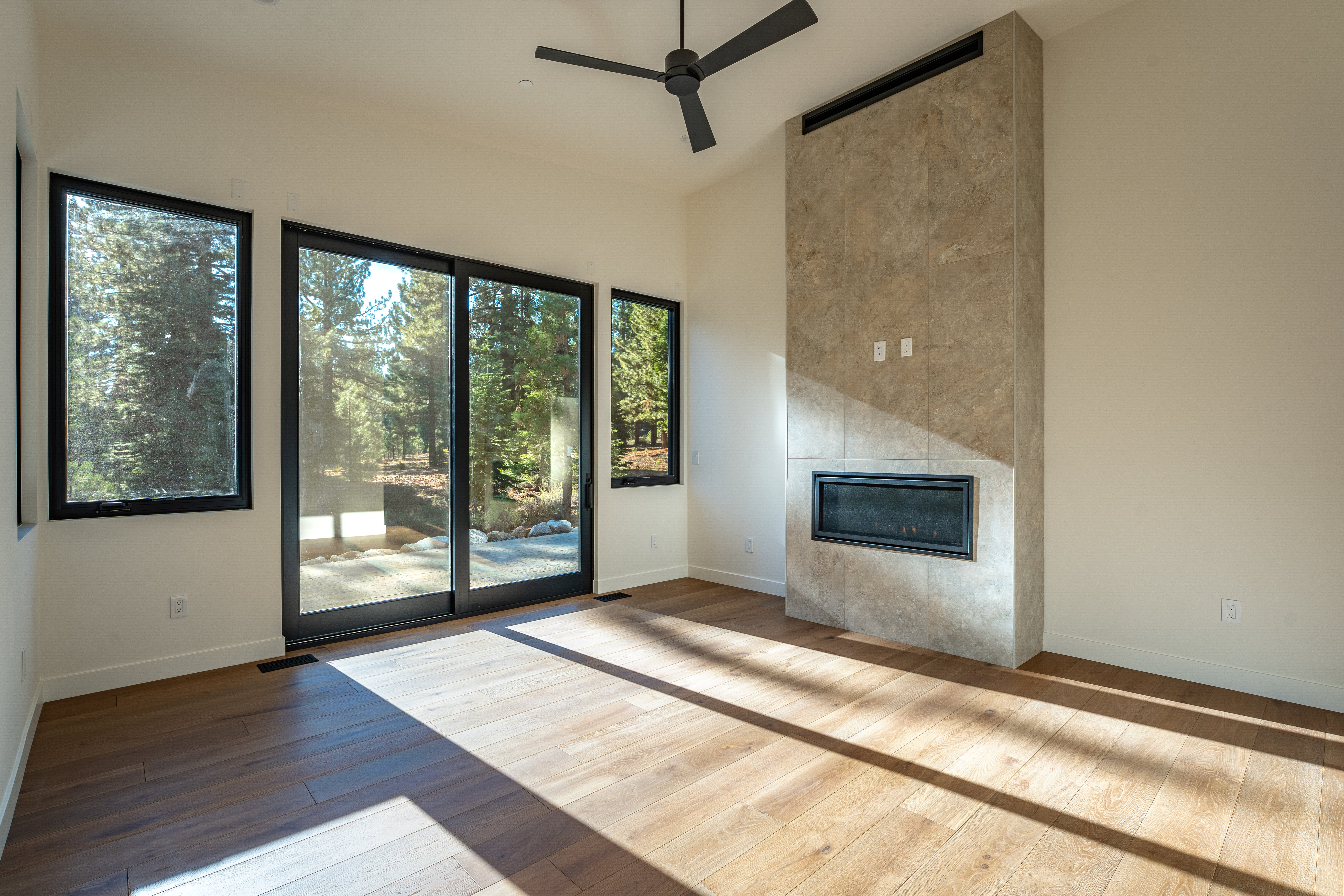 This is a bright and modern living room featuring hardwood floors and a contemporary fireplace with a stone surround. Large windows and a sliding glass door offer views of the surrounding trees and landscape, bringing natural light into the space. A ceiling fan adds to the room's comfort and style.