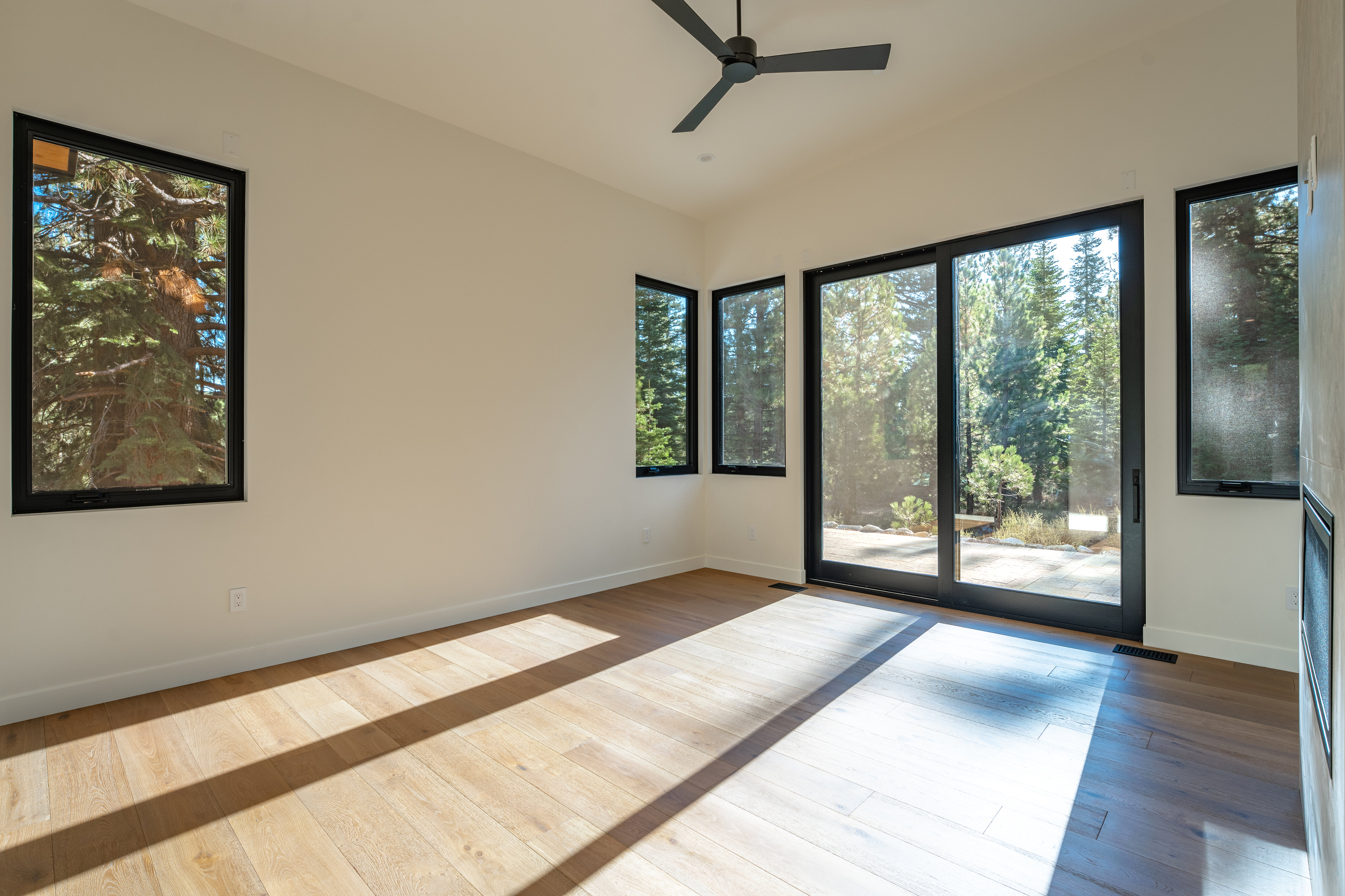This is a bright and airy primary bedroom featuring light hardwood floors and large windows that offer views of the surrounding trees. The room is minimalist in style, with a ceiling fan and neutral-colored walls. The abundance of natural light creates a serene and inviting atmosphere.