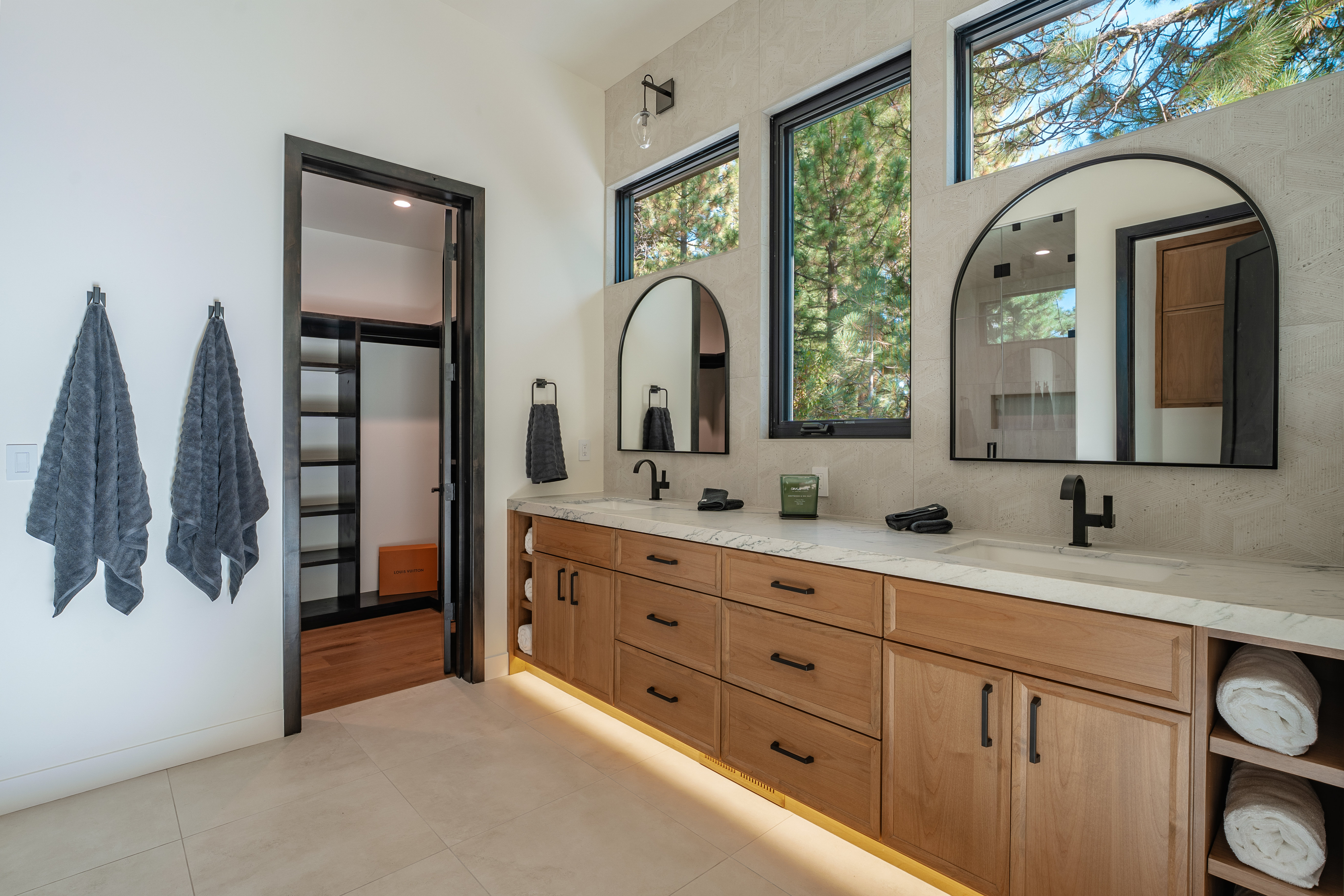 This is a primary bathroom featuring a double vanity with light wood cabinetry, marble countertops, and black hardware. Arched mirrors are positioned above each sink, complemented by black-framed windows that offer natural light and views of the outdoors. The space includes a walk-in closet and is decorated with gray towels, creating a modern and luxurious feel.