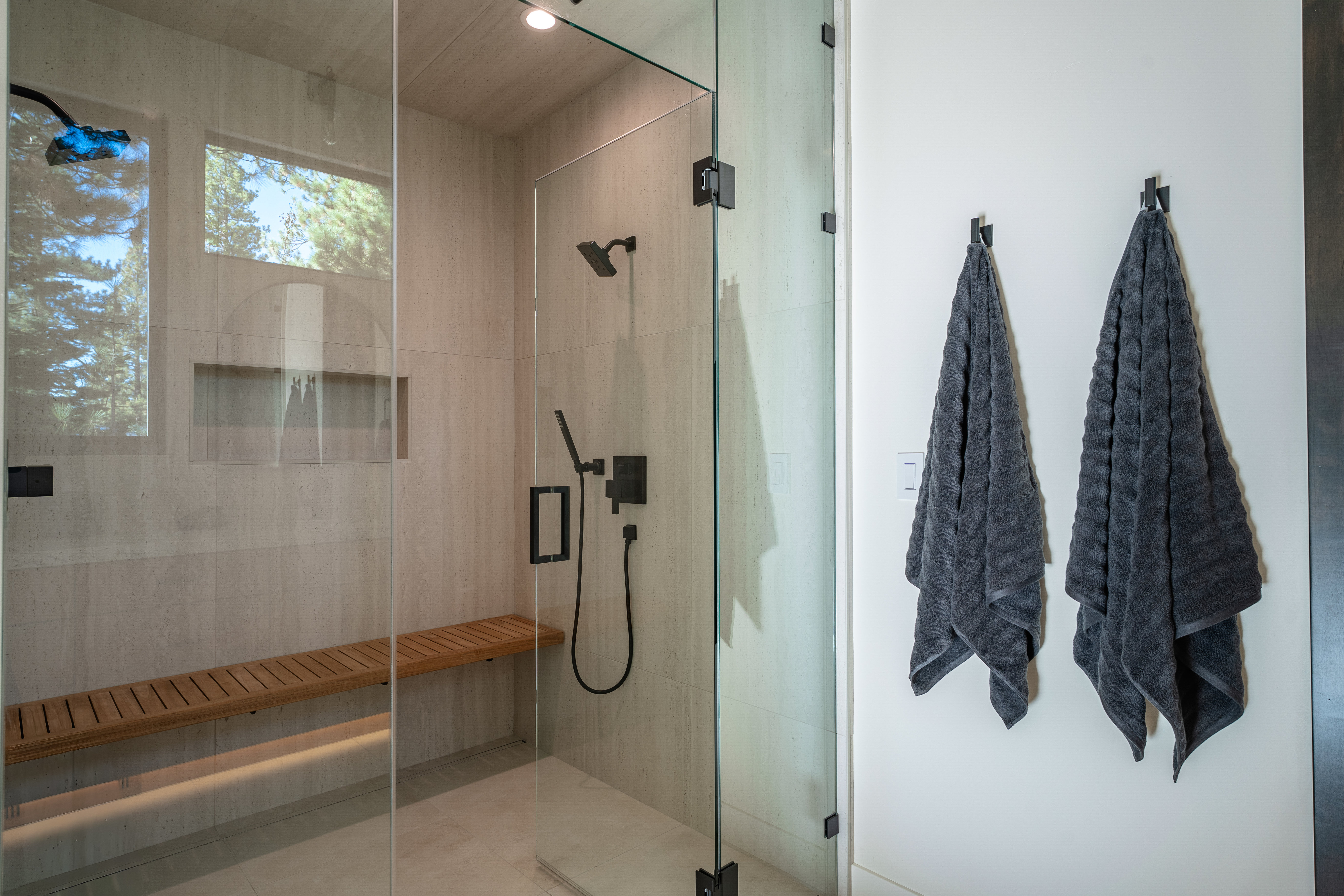 This is a modern primary bathroom featuring a glass-enclosed shower with a built-in wooden bench and a rainfall showerhead. The walls are tiled in a neutral tone, and natural light streams in through a window. Two dark gray towels hang on hooks on the adjacent wall, adding a touch of contrast.