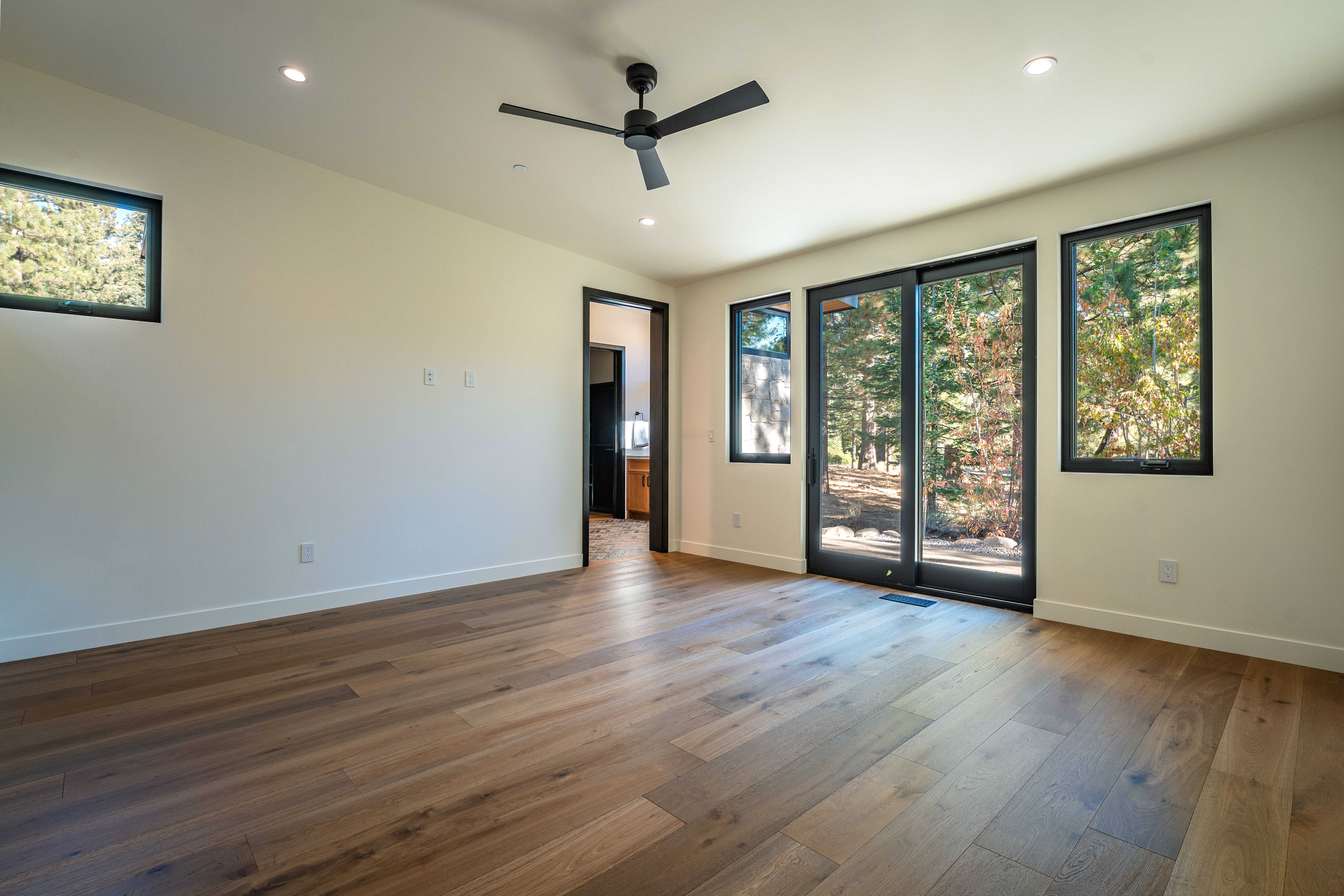 This is a spacious primary bedroom featuring light-colored walls, hardwood flooring, and a modern ceiling fan. Natural light floods the room through multiple windows and a sliding glass door, offering views of the surrounding trees. The room has a minimalist aesthetic with clean lines and a neutral color palette.