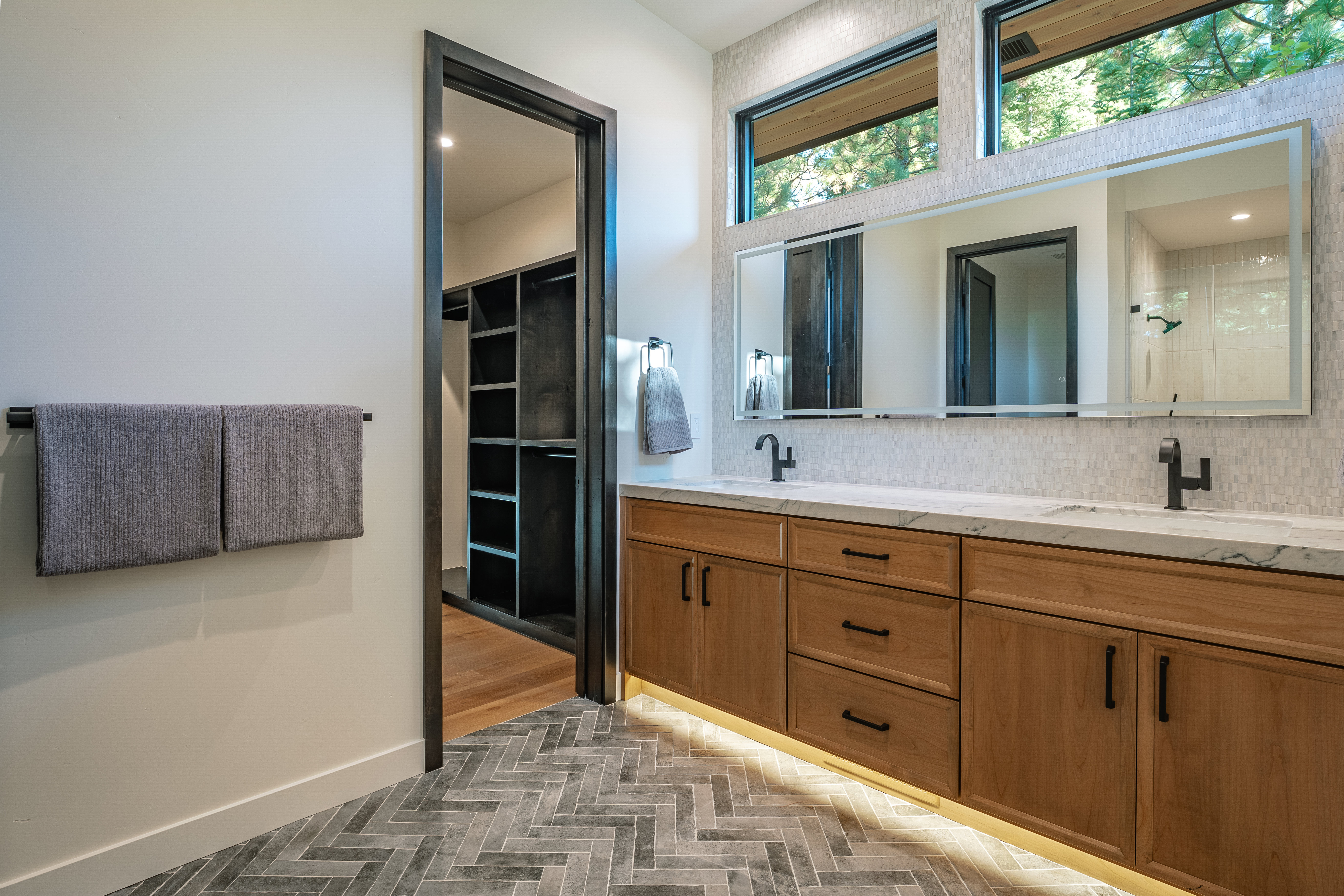 This is a well-lit primary bathroom featuring a double vanity with wood cabinetry, marble countertops, and modern black fixtures. A large mirror spans the length of the vanity, reflecting the natural light from the clerestory windows above. The floor is tiled in a herringbone pattern, and a doorway leads to a walk-in closet, creating a luxurious and functional space.