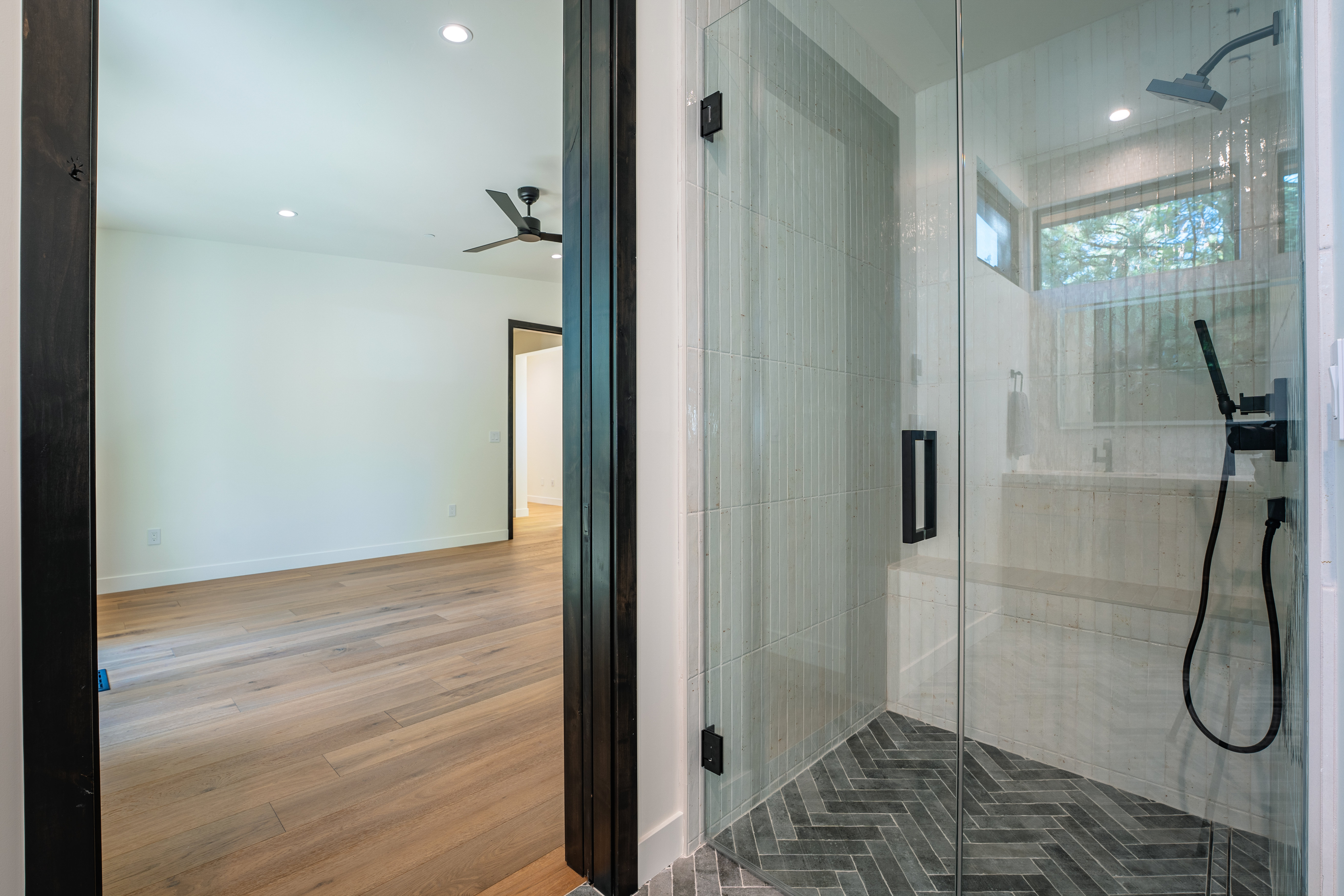 This image showcases a modern primary bathroom, focusing on the glass-enclosed shower with stylish gray vertical tiles and dark herringbone floor tiles. The shower features matte black fixtures, including a shower head and handheld sprayer. Through the doorway, a glimpse of the bedroom reveals hardwood floors and white walls, creating a bright and airy atmosphere.
