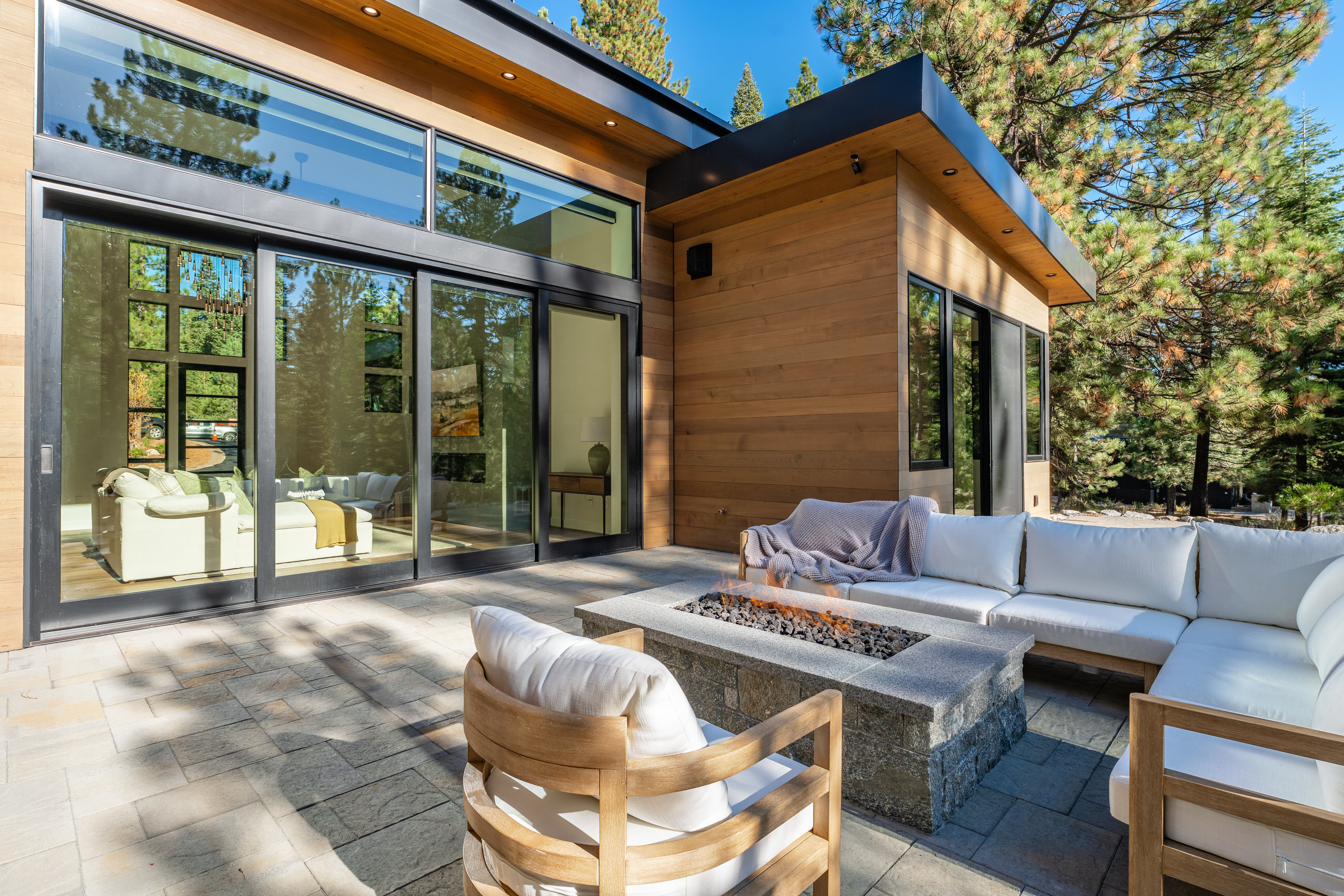 This image showcases a modern patio area featuring a stone fire pit surrounded by comfortable outdoor seating. The patio is paved with large stone tiles, and the backdrop includes a contemporary home with large windows and wood siding. The scene is bathed in natural light, creating an inviting and luxurious outdoor living space.