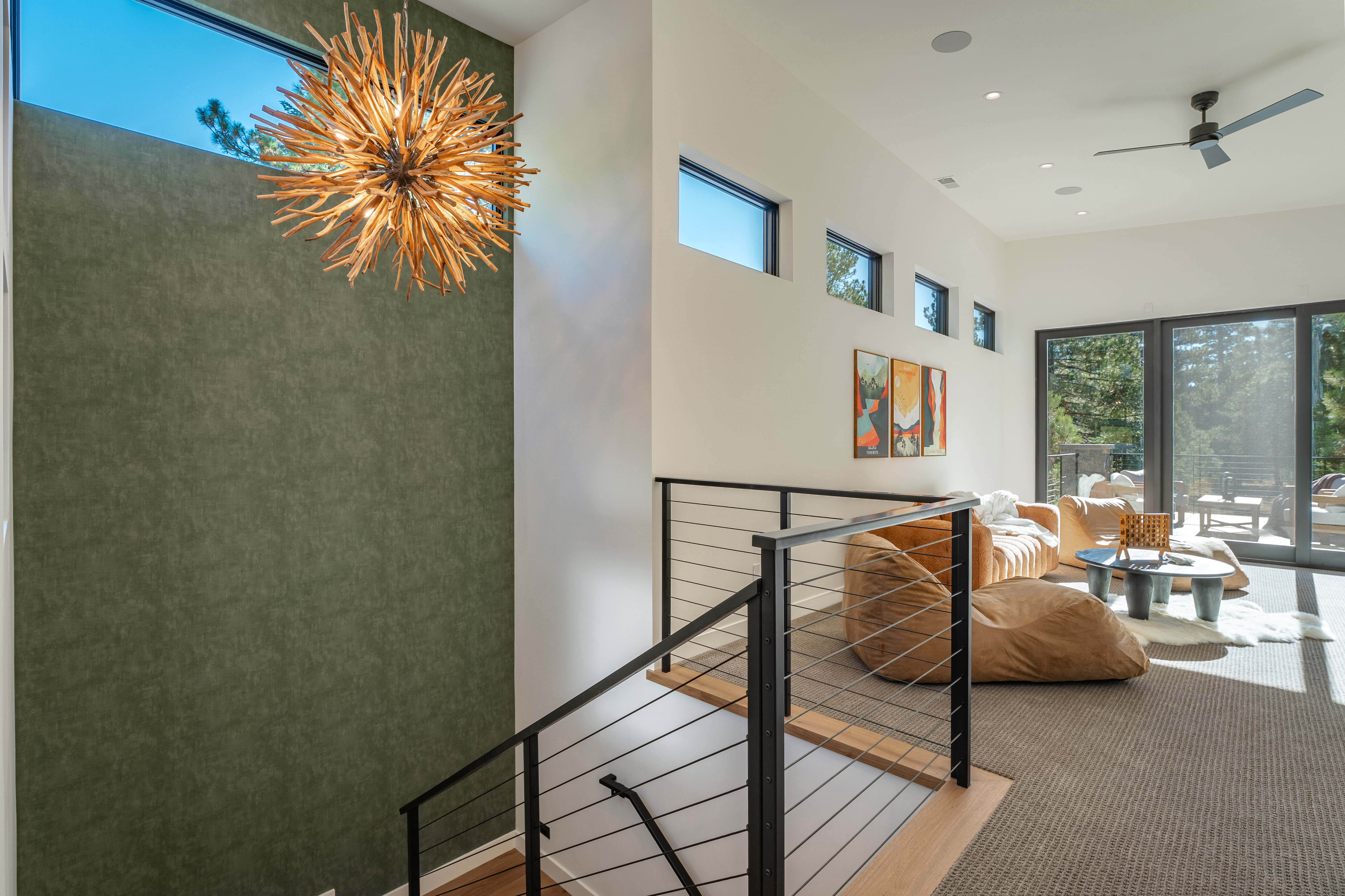 This interior shot showcases a modern hallway and staircase. The staircase features wooden steps with a sleek black metal railing. A striking, spherical chandelier hangs near a textured green wall, while the hallway leads to a bright living area with comfortable seating and access to an outdoor deck through large sliding glass doors.