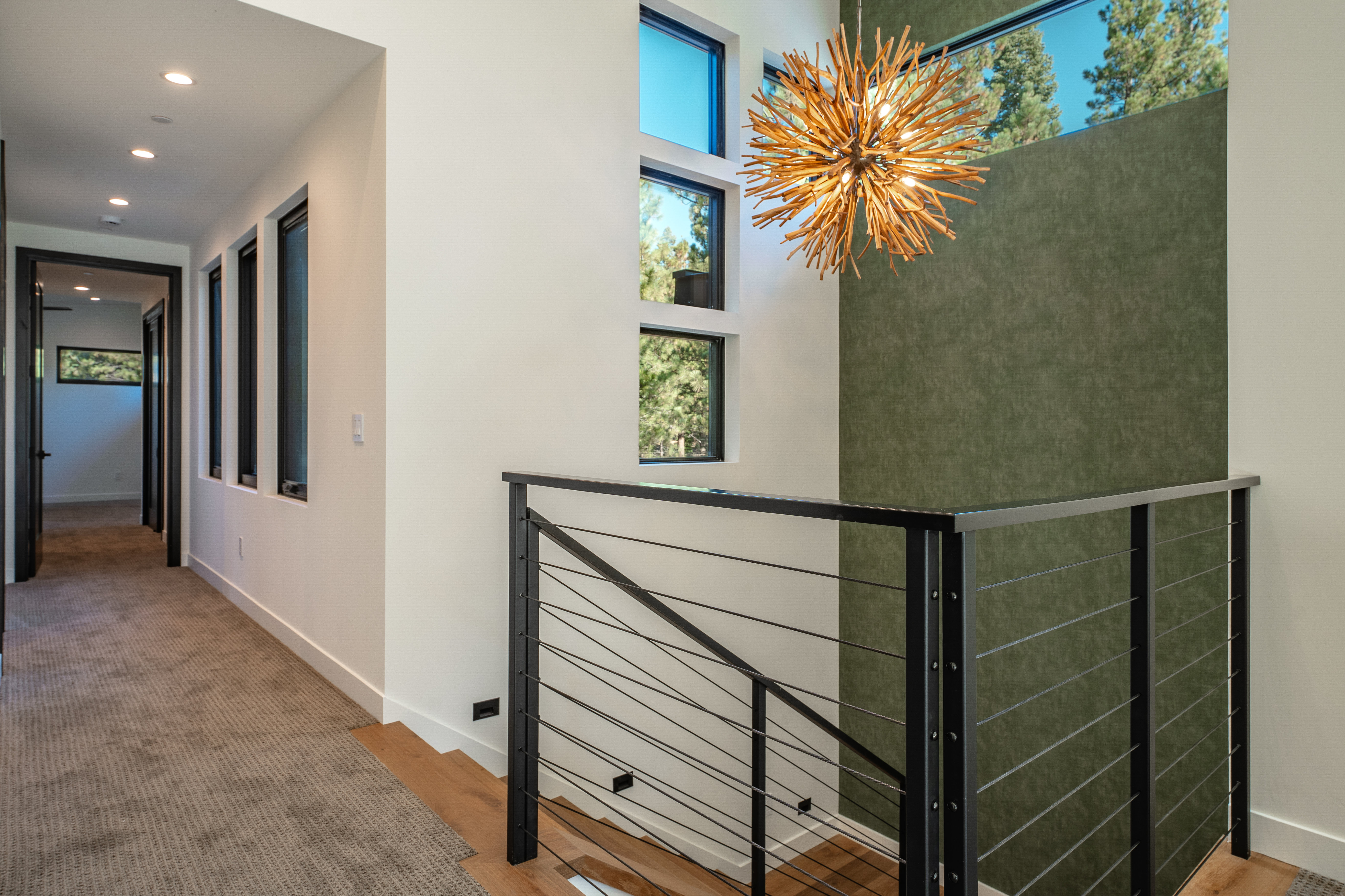 This interior shot showcases a modern hallway and staircase area. The hallway features a neutral-toned carpet, recessed lighting, and a series of narrow, vertical windows. The staircase is defined by a sleek black railing with horizontal cable wires, complemented by wooden steps and a unique, spherical chandelier hanging above, adding a touch of artistic flair to the space.