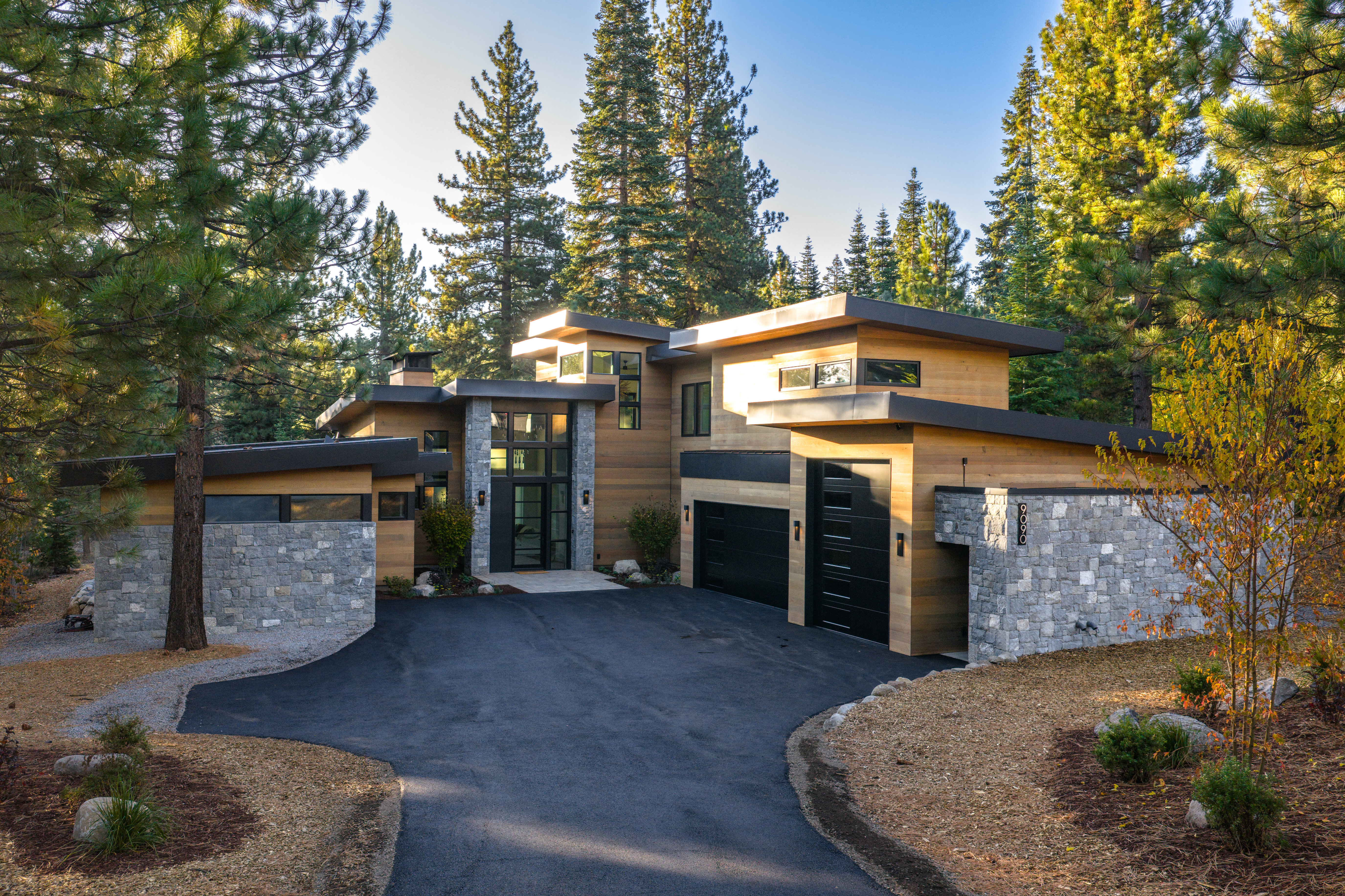 This is a front exterior view of a modern, two-story home with a flat roof and a combination of wood and stone facade. The house features a paved driveway leading to a two-car garage with black doors. Tall pine trees surround the property, enhancing its privacy and natural setting.