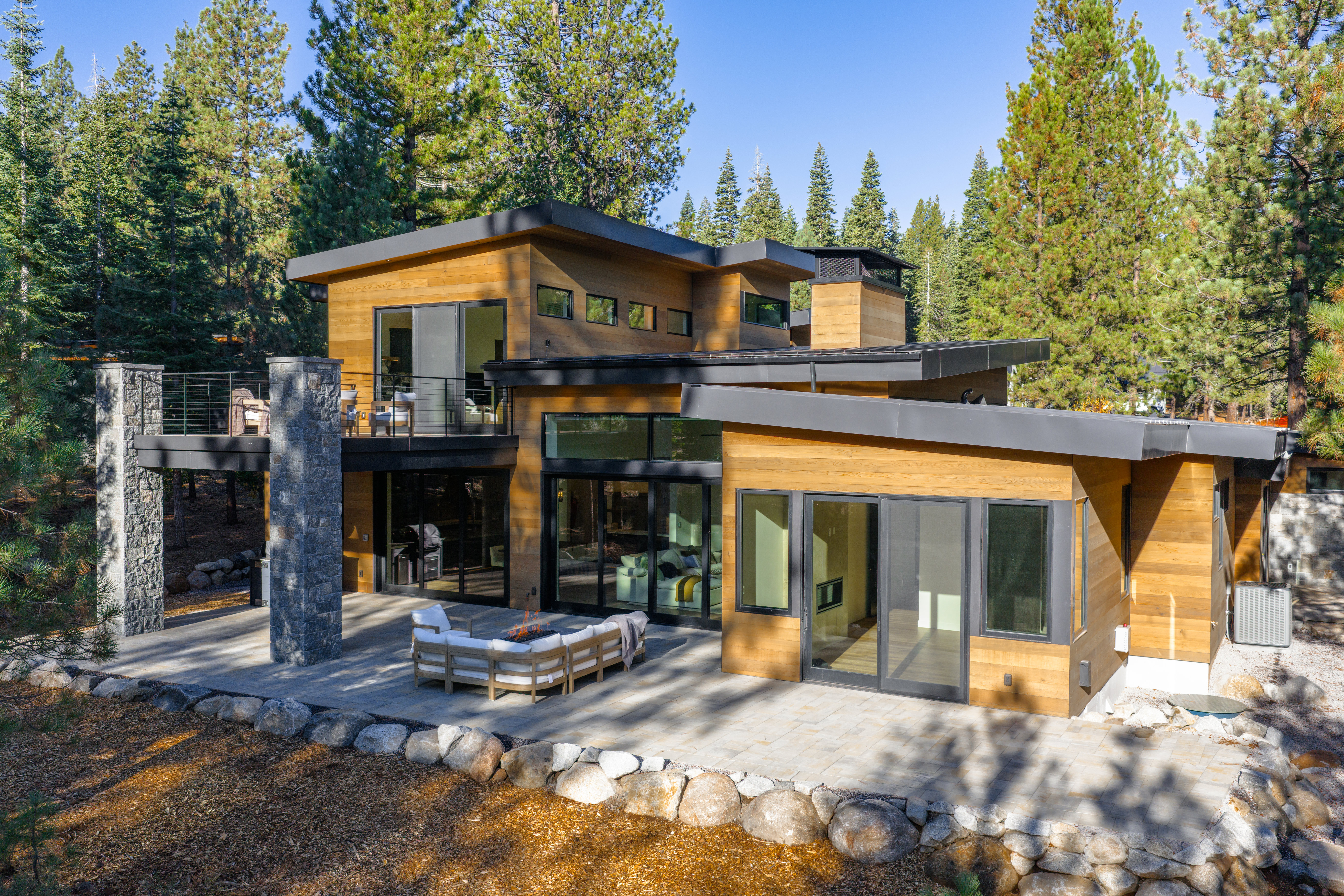 This is a rear exterior view of a modern home, showcasing its architectural design and outdoor living space. The house features wood siding, large windows, and a flat roof with dark trim. A stone patio with a fire pit and outdoor seating area extends from the house, surrounded by landscaping and mature trees, creating a serene and inviting atmosphere.