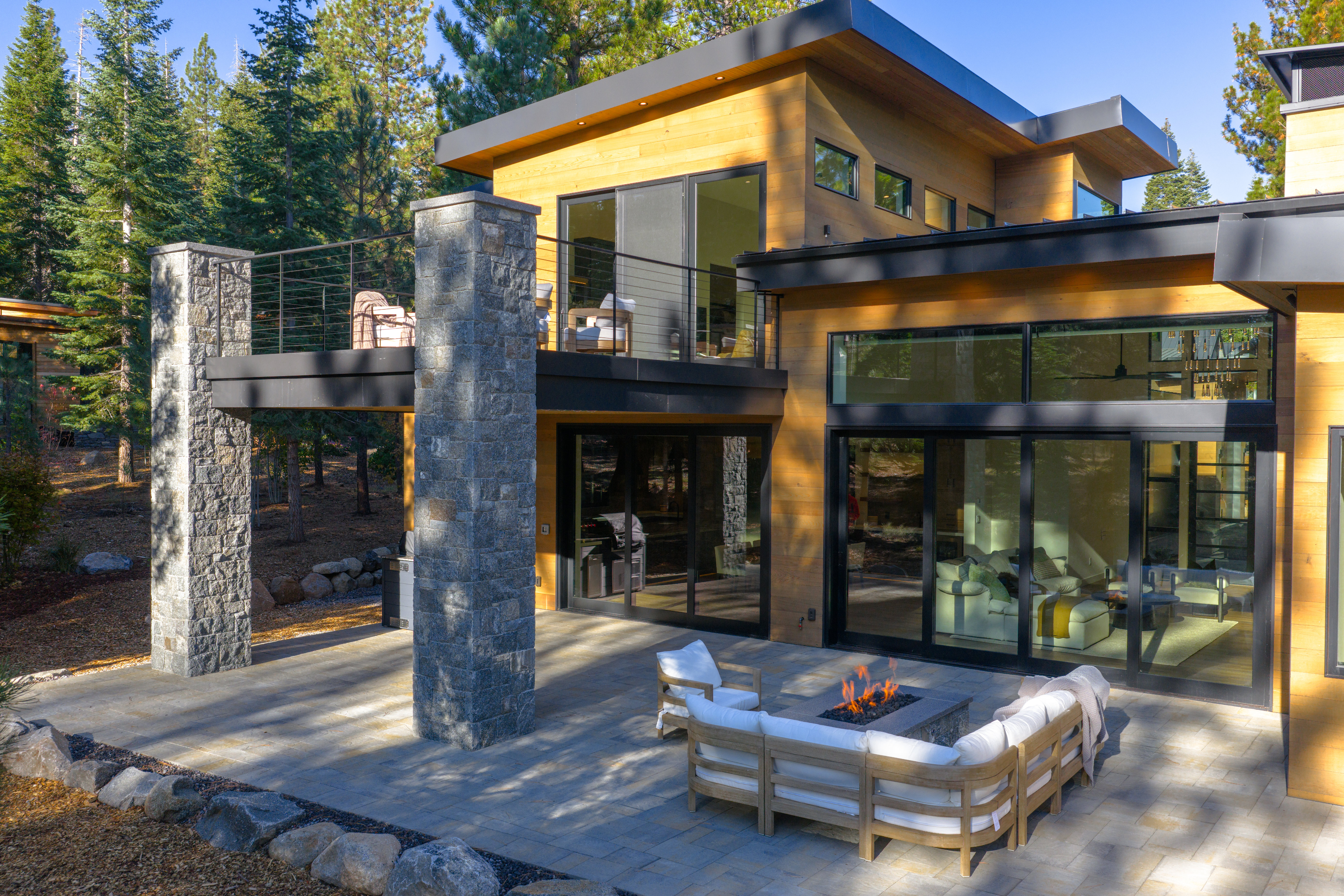 This image showcases a modern home's outdoor living space, featuring a spacious patio with a fire pit and comfortable seating. The architecture is characterized by clean lines, wood siding, and large windows that offer views into the interior. A balcony with stone pillars adds architectural interest and provides an elevated outdoor area.