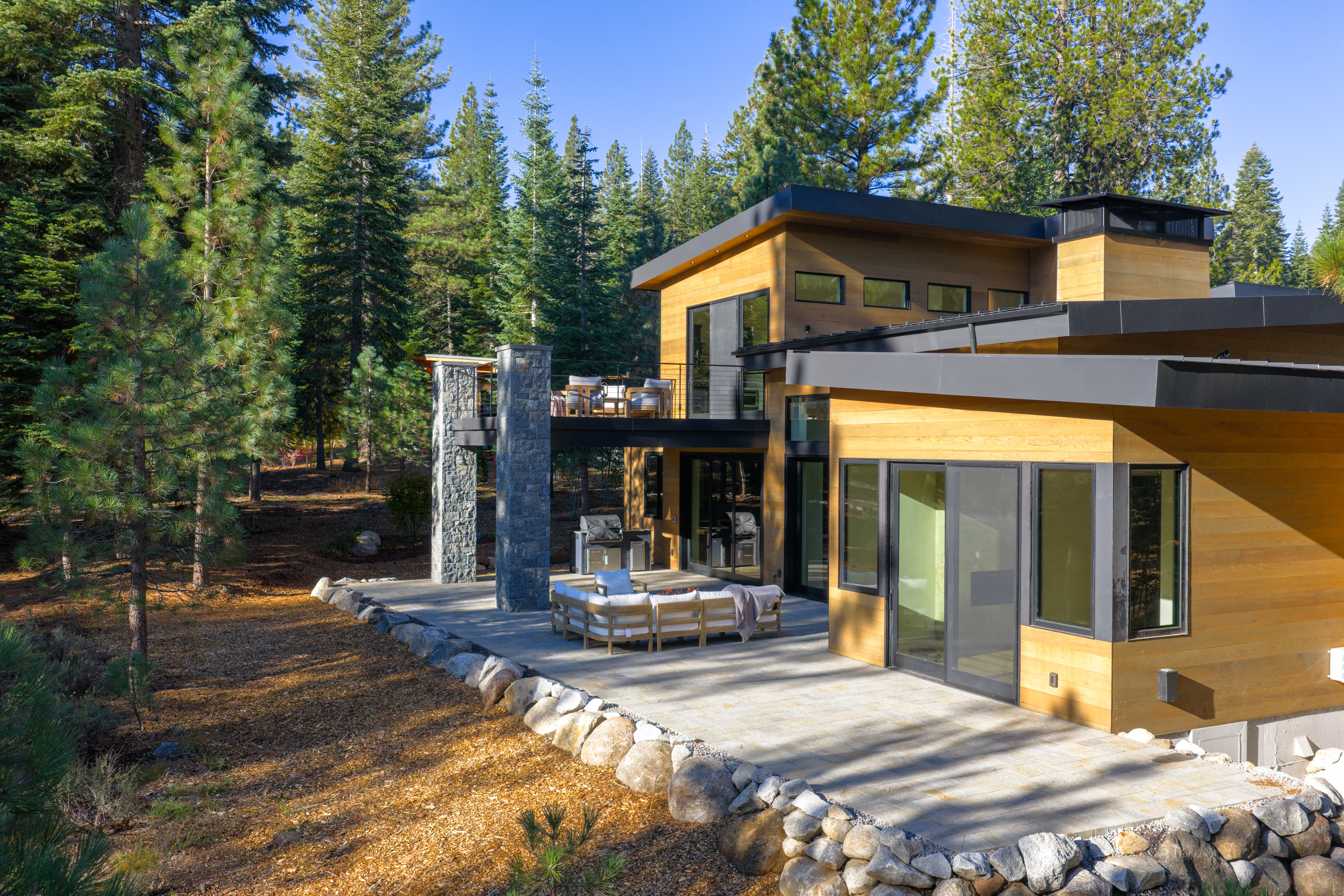 This is a rear exterior view of a modern home, showcasing a spacious patio area with outdoor seating and a grill. The house features wood siding, black-framed windows, and a second-story balcony. The surrounding landscape includes mature trees and a rock border, creating a private and serene setting.