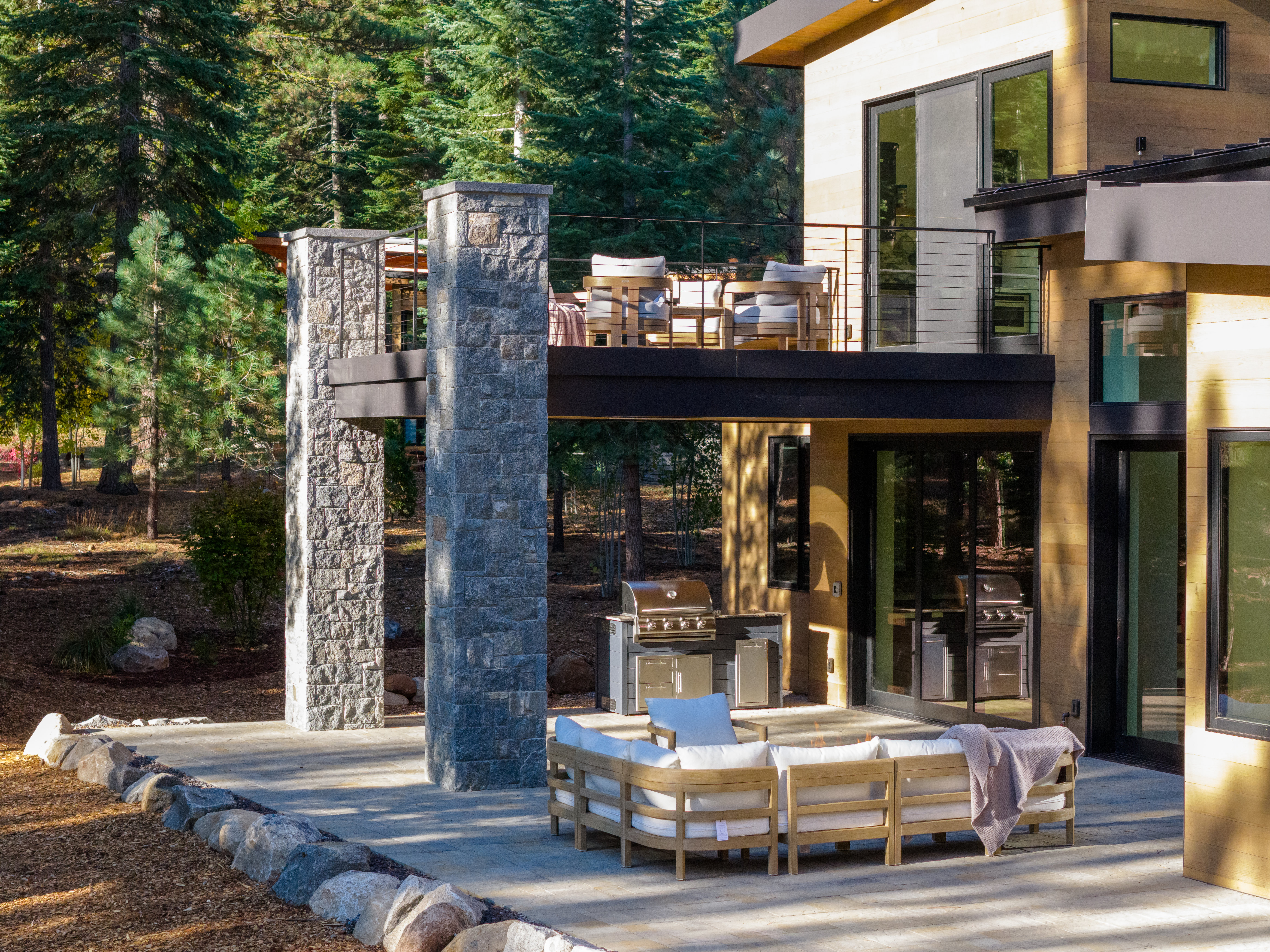 This image showcases a modern home's outdoor living space, featuring a spacious patio with a comfortable seating area and a built-in grilling station. A second-story balcony with outdoor seating overlooks the patio, supported by stone pillars that add architectural interest. The surrounding landscape includes mature trees, creating a private and serene atmosphere, perfect for relaxation and entertaining.