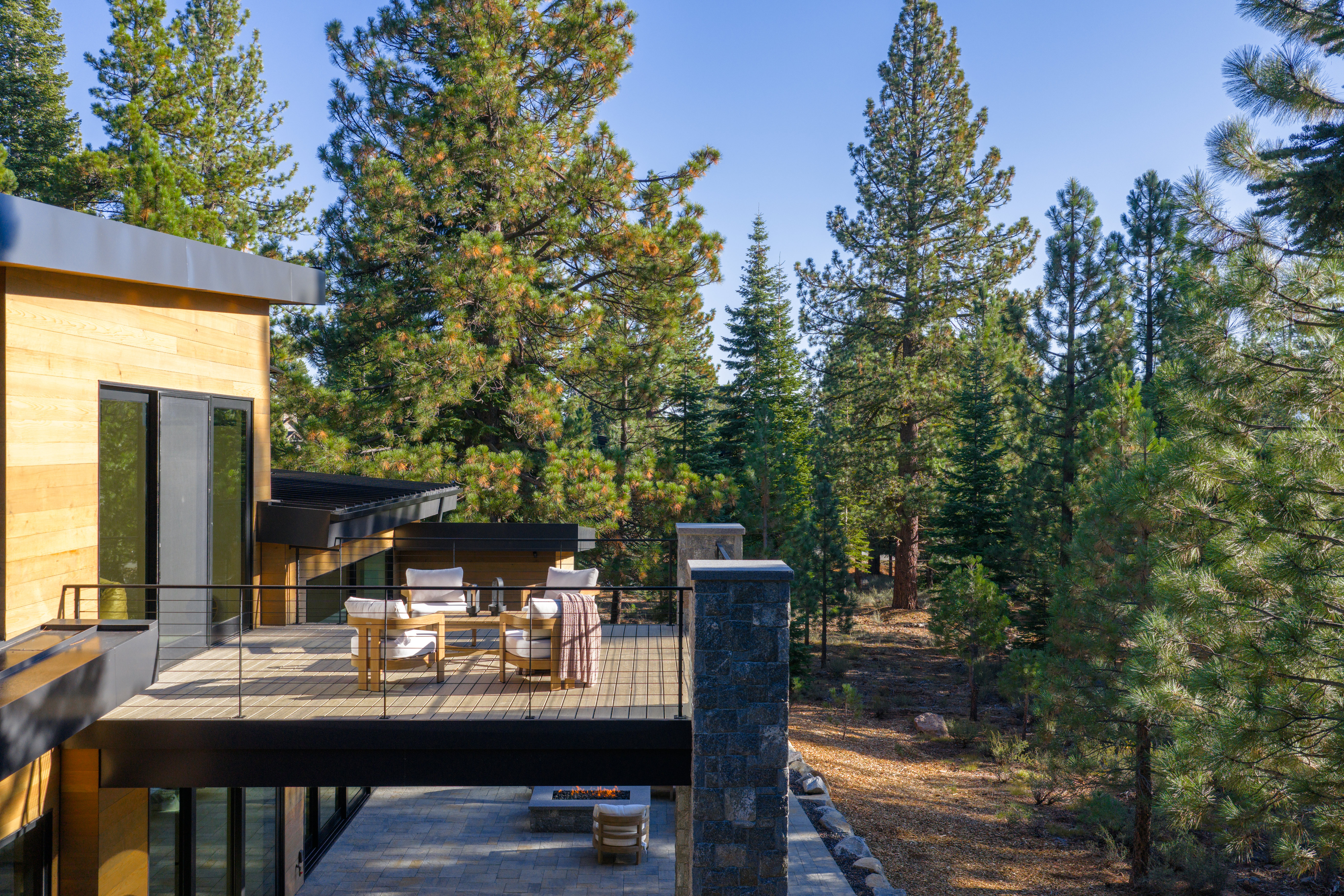 This image showcases a modern home's deck or balcony, featuring stylish outdoor furniture arranged for relaxation and conversation. The deck is constructed with light-toned wood and has a sleek black railing, offering views of the surrounding lush forest. Below the deck is a stone patio with a fire pit, creating an inviting outdoor living space.