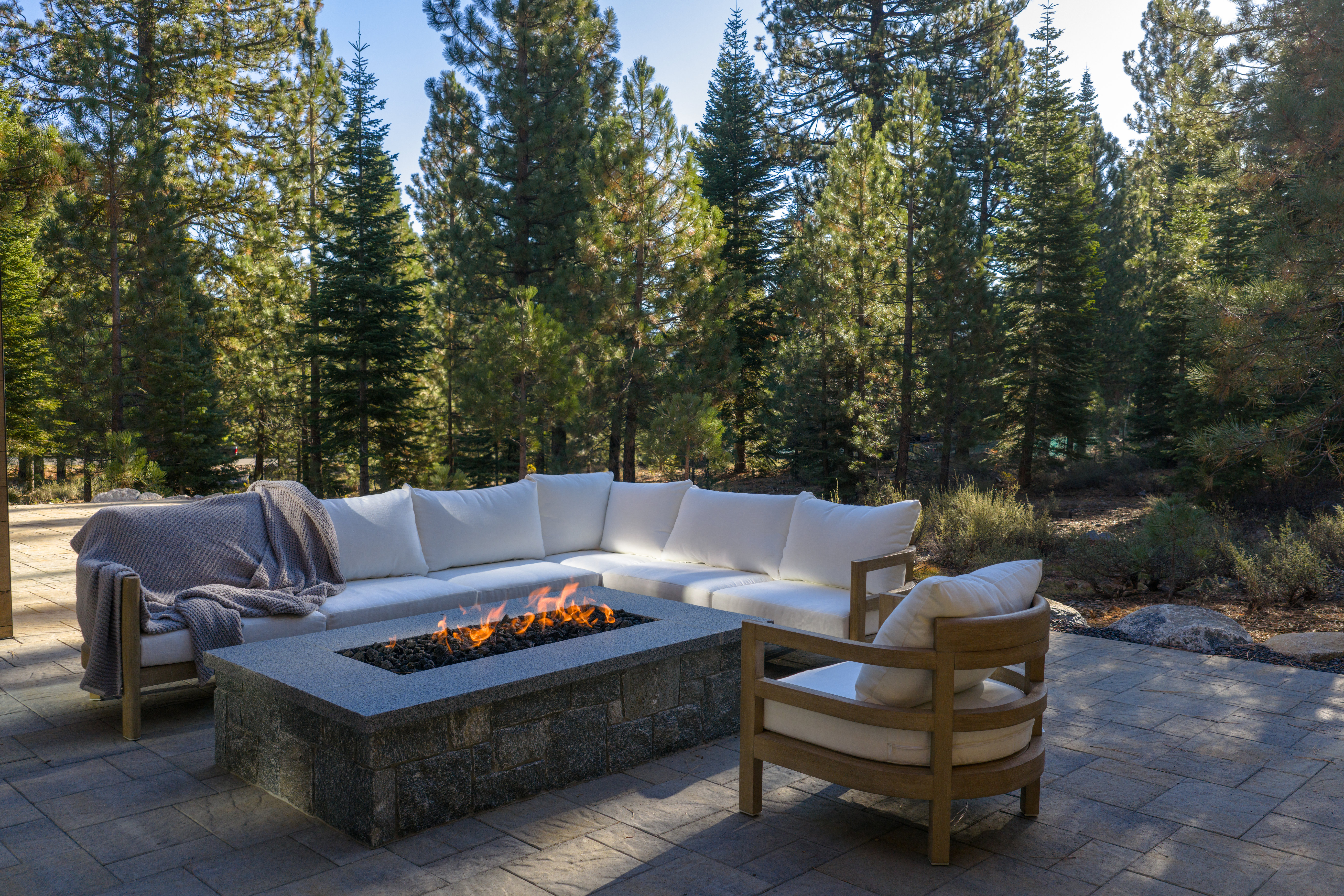 This image showcases a luxurious outdoor patio area, featuring a large sectional sofa with white cushions and a modern fire pit with visible flames. A comfortable armchair complements the seating arrangement, all set on a paved patio surrounded by tall evergreen trees. The scene evokes a sense of relaxation and upscale outdoor living.