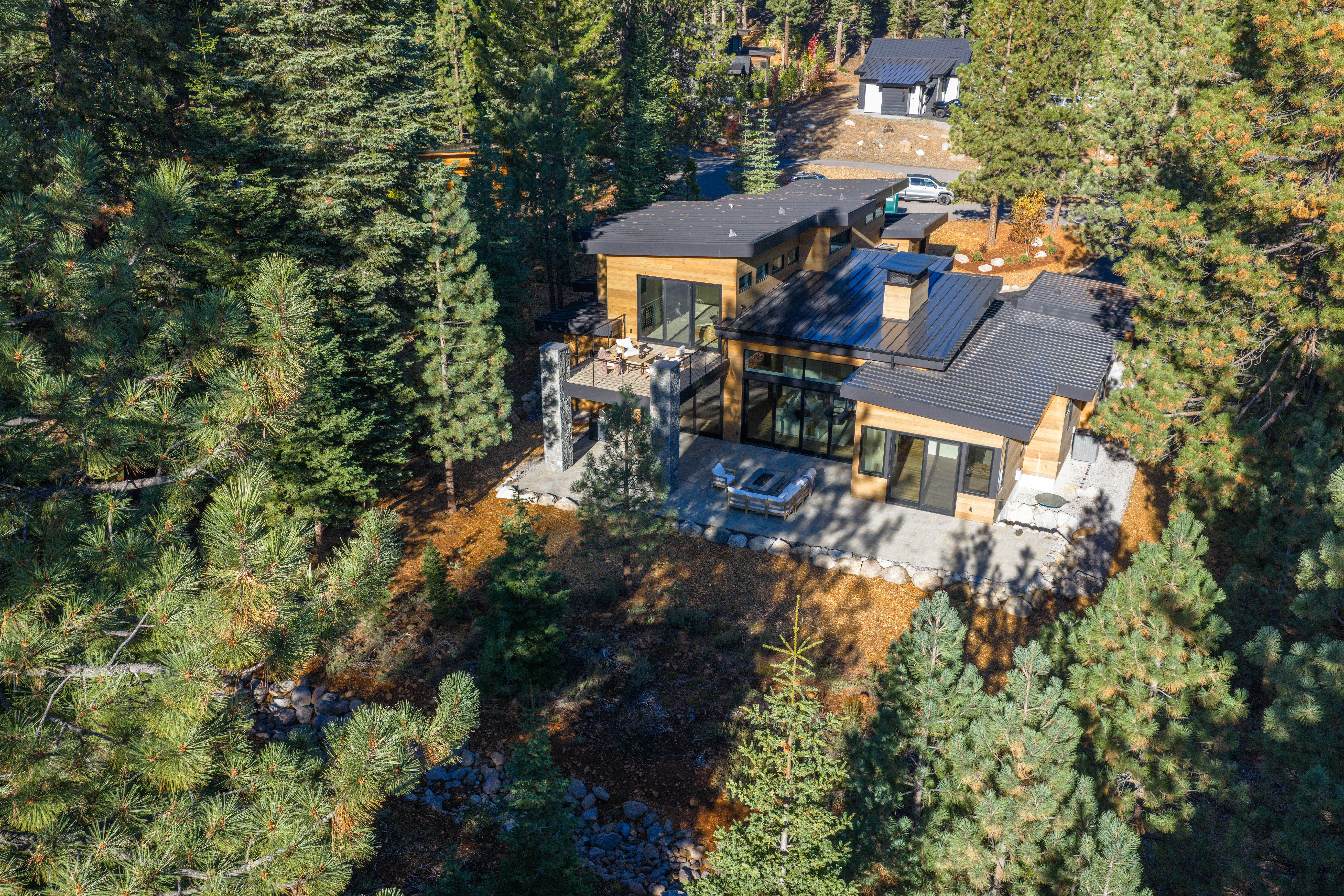 This aerial shot showcases a modern home nestled among tall pine trees. The house features a combination of wood siding and dark metal roofing, with a spacious patio area including a fire pit and outdoor seating. The surrounding landscape is a mix of mature trees and natural ground cover, creating a sense of privacy and seclusion.