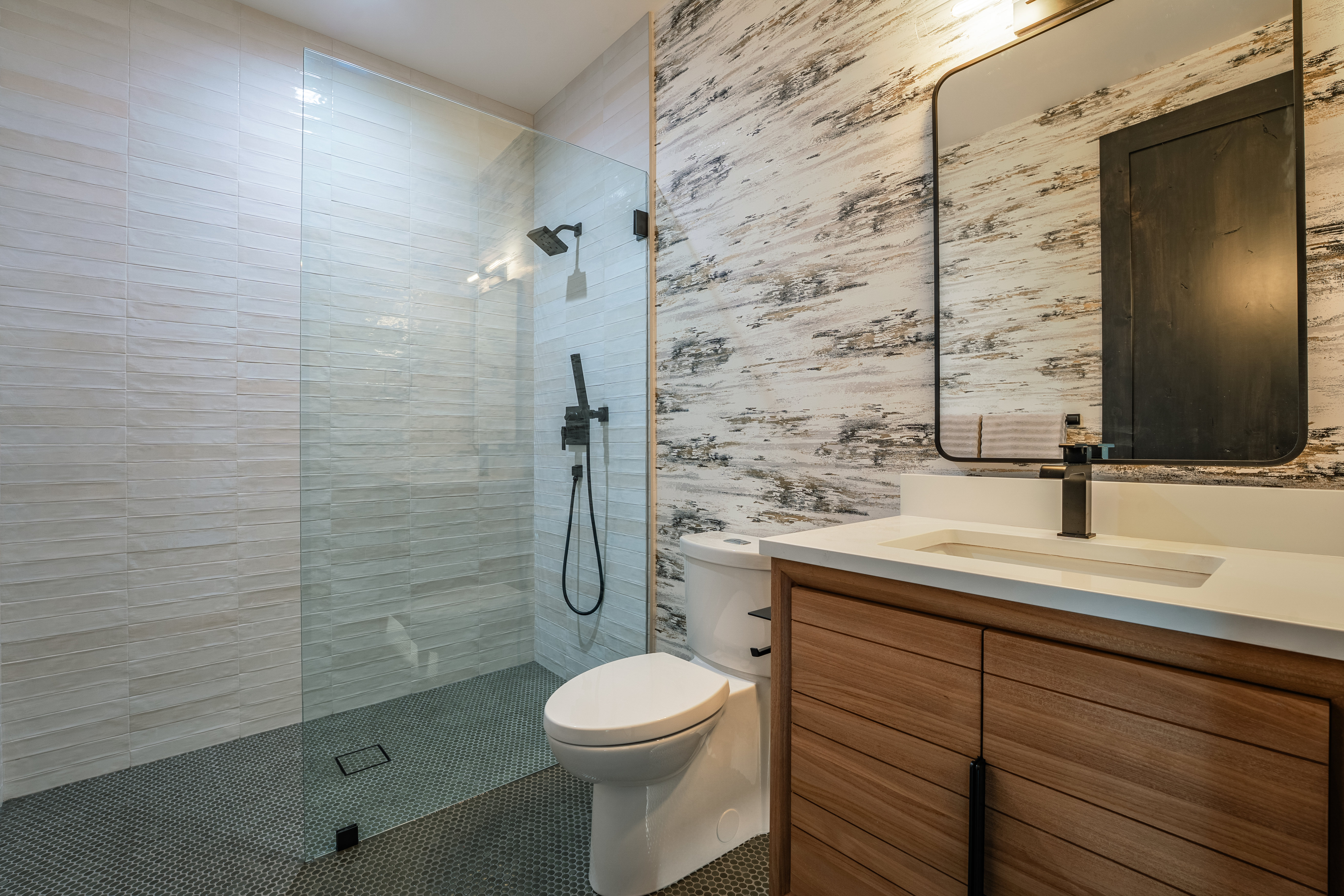 This is a modern bathroom featuring a glass-enclosed shower with black fixtures, a toilet, and a vanity with a light wood cabinet and white countertop. The walls are tiled with a combination of textured white tiles in the shower and a decorative, horizontally-striped pattern on the adjacent wall. The floor is covered in small, dark gray hexagonal tiles, creating a sleek and contemporary look.