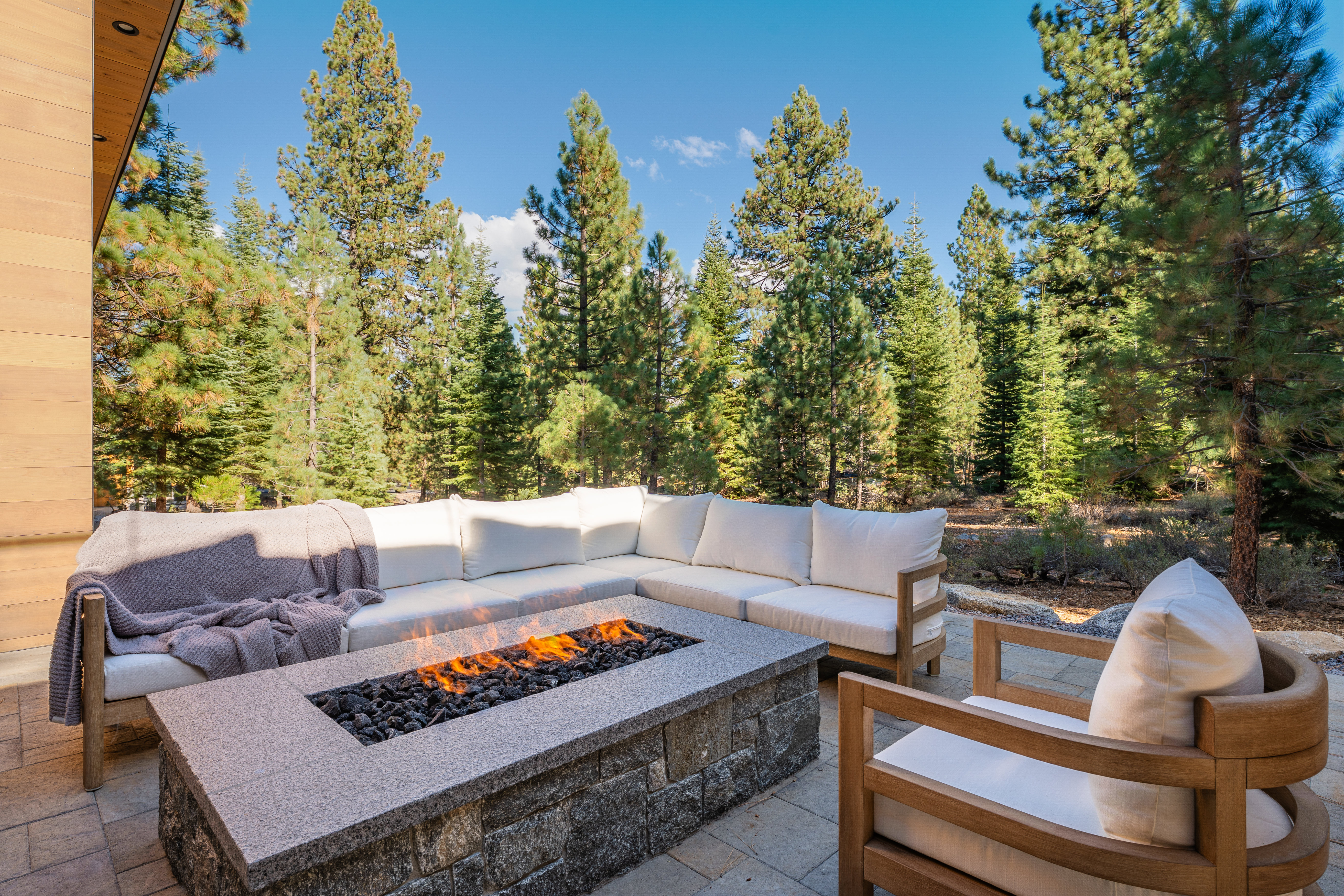 This image showcases a luxurious outdoor patio area, complete with a large, comfortable sectional sofa and a modern fire pit. The seating area is surrounded by tall pine trees, creating a serene and private atmosphere. A stylish armchair complements the sofa, enhancing the inviting ambiance of this outdoor living space.