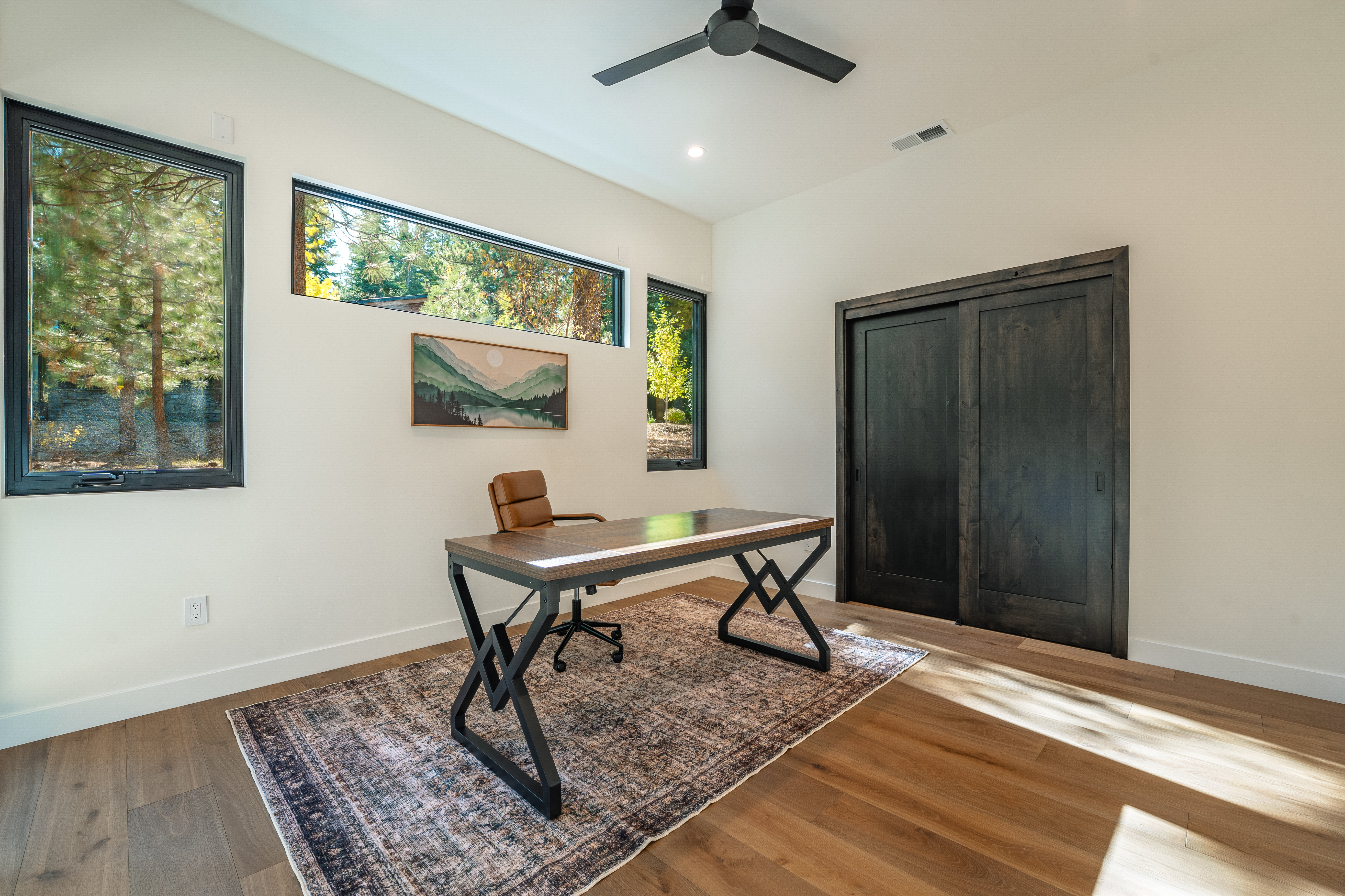 This is an interior shot of a modern office space. The room features a wooden desk with black metal legs, a brown leather office chair, and a patterned rug on hardwood floors. Natural light streams in through several windows, and a sliding barn door is visible on the right side of the frame.