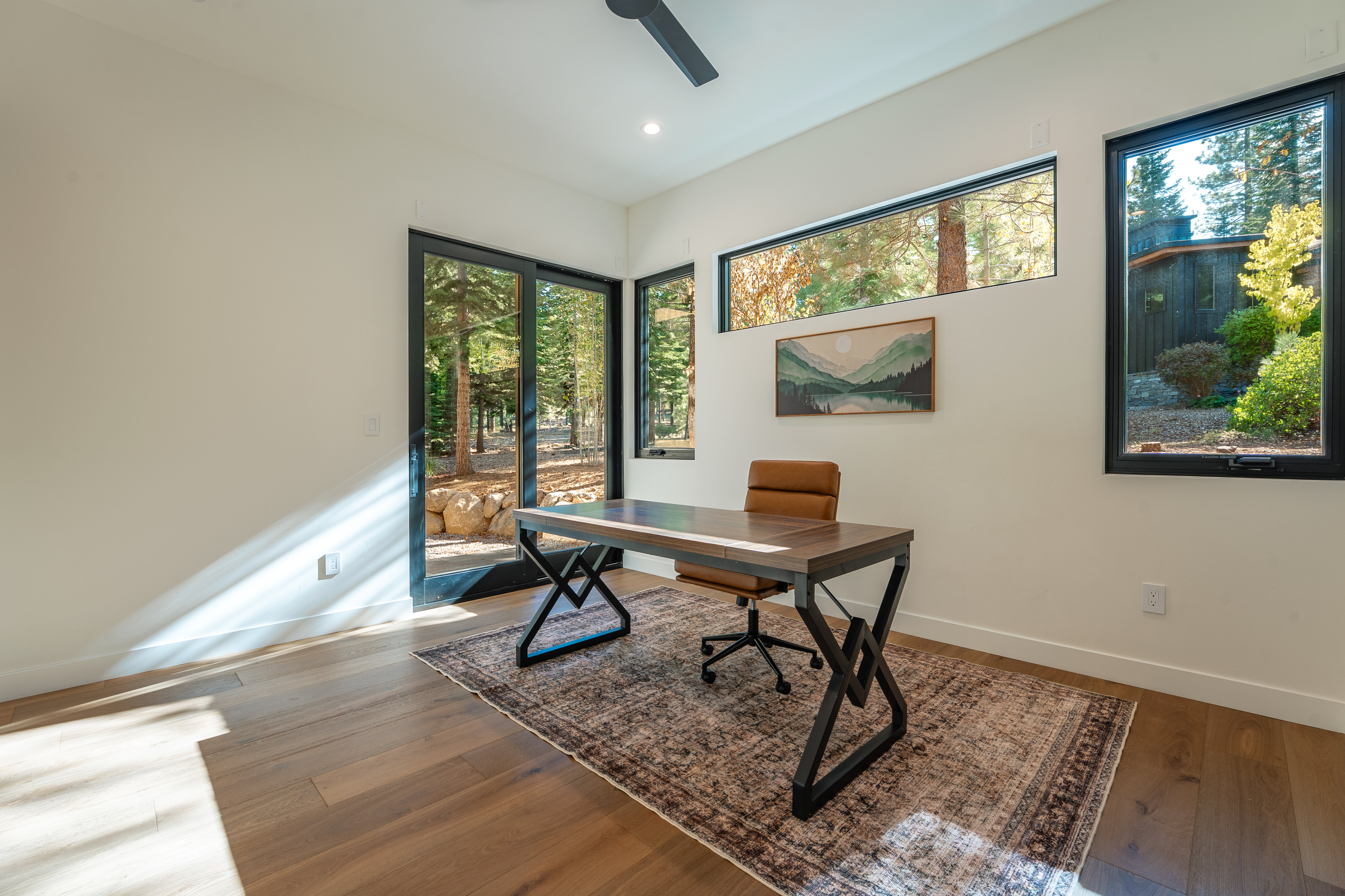 This is an interior shot of a modern office space featuring a wooden desk with a black metal frame, a brown leather office chair, and a patterned rug. Large windows provide natural light and views of the surrounding trees, enhancing the connection to nature. The room has a minimalist aesthetic with white walls and hardwood floors, creating a clean and inviting workspace.