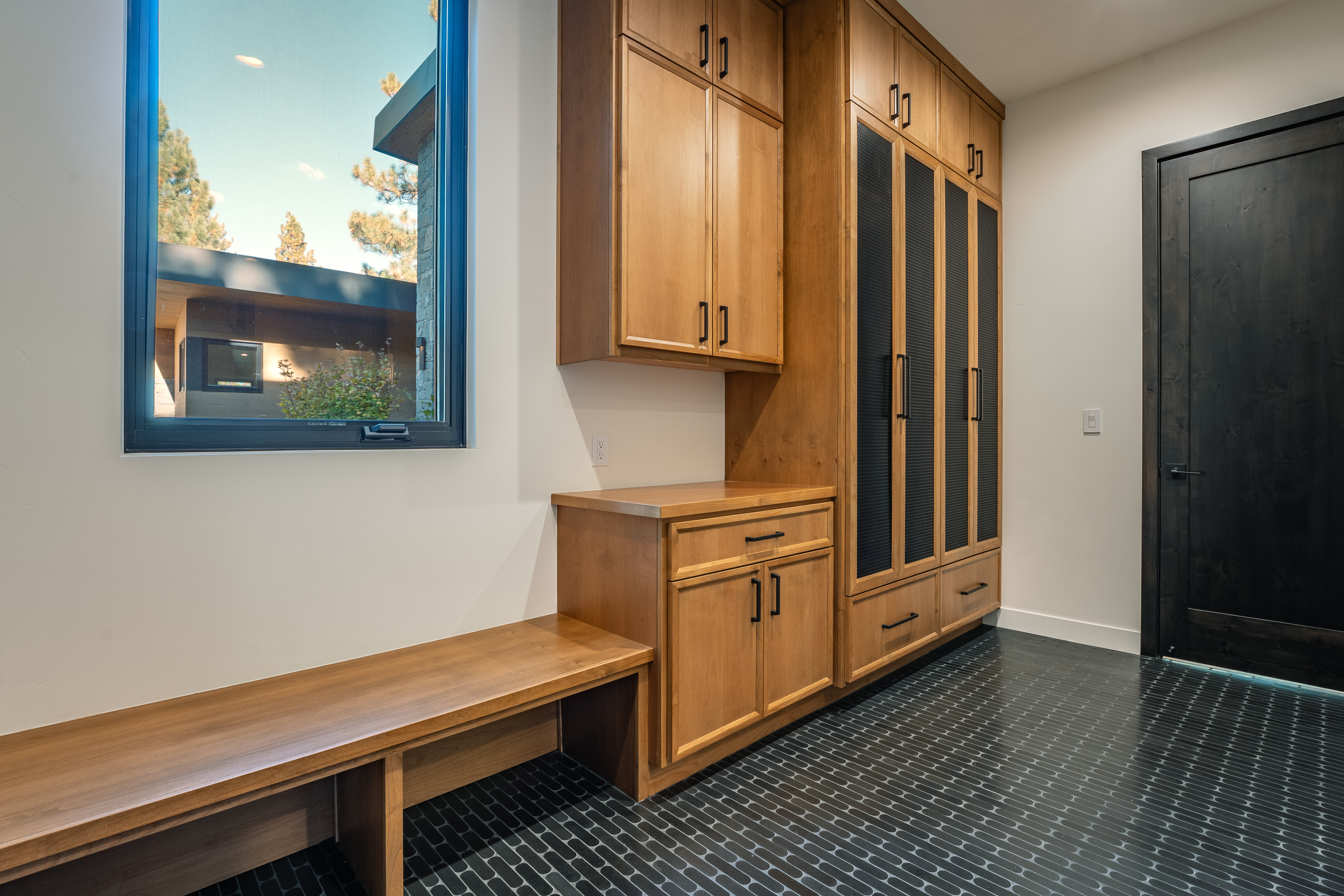 This interior shot showcases a well-organized laundry room with custom wooden cabinetry and black hardware. A built-in bench sits beneath a window, providing a convenient spot to sit. The black tiled floor adds a touch of modern elegance to the space, while the dark door on the right provides a striking contrast.