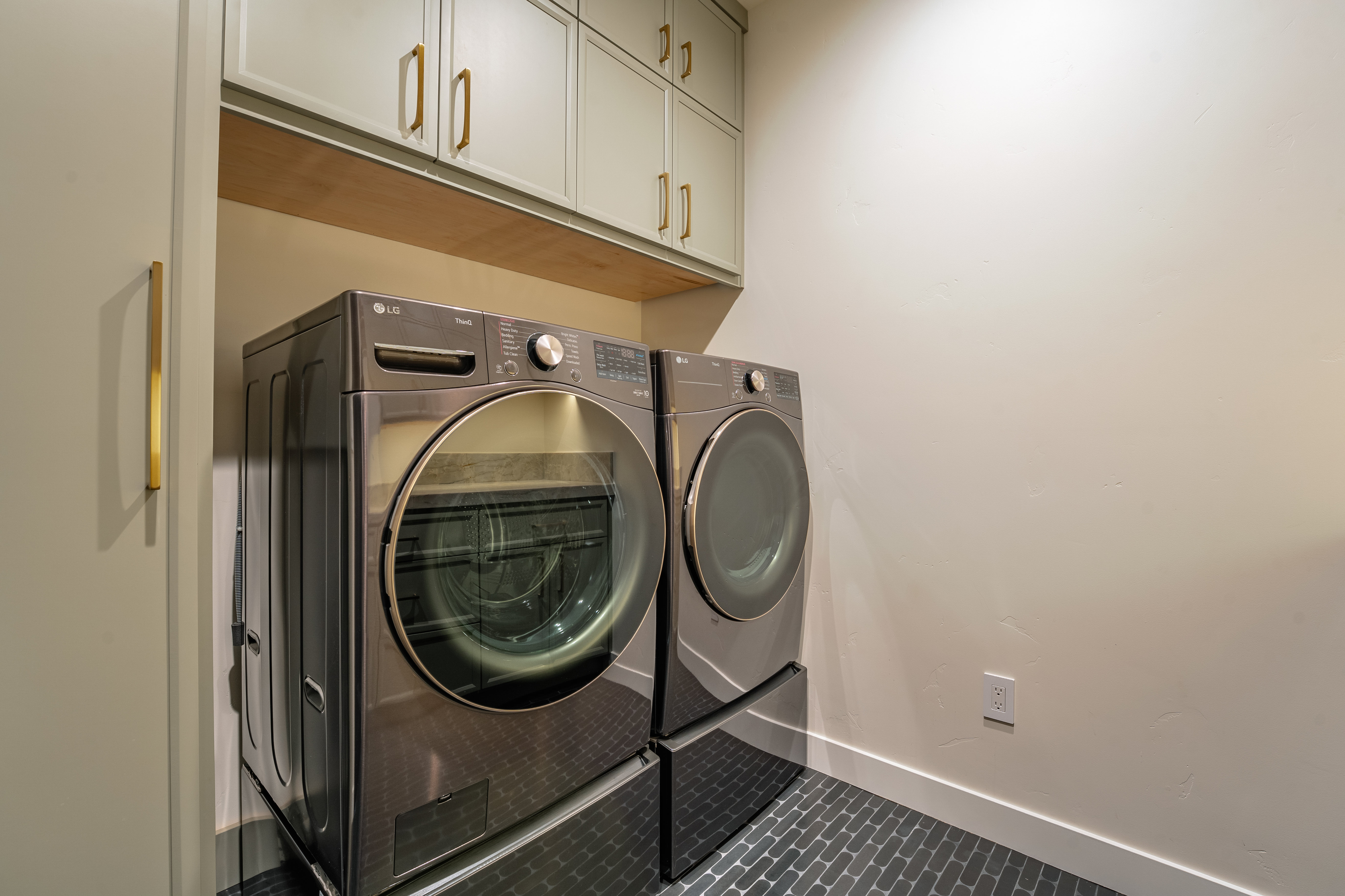 This is a well-organized laundry room featuring a modern LG washer and dryer set in a dark metallic finish. The units are placed on a dark, patterned tile floor, and overhead cabinets in a muted green provide ample storage. The room is clean and functional, with a neutral wall color and white trim, creating a tidy and efficient space.