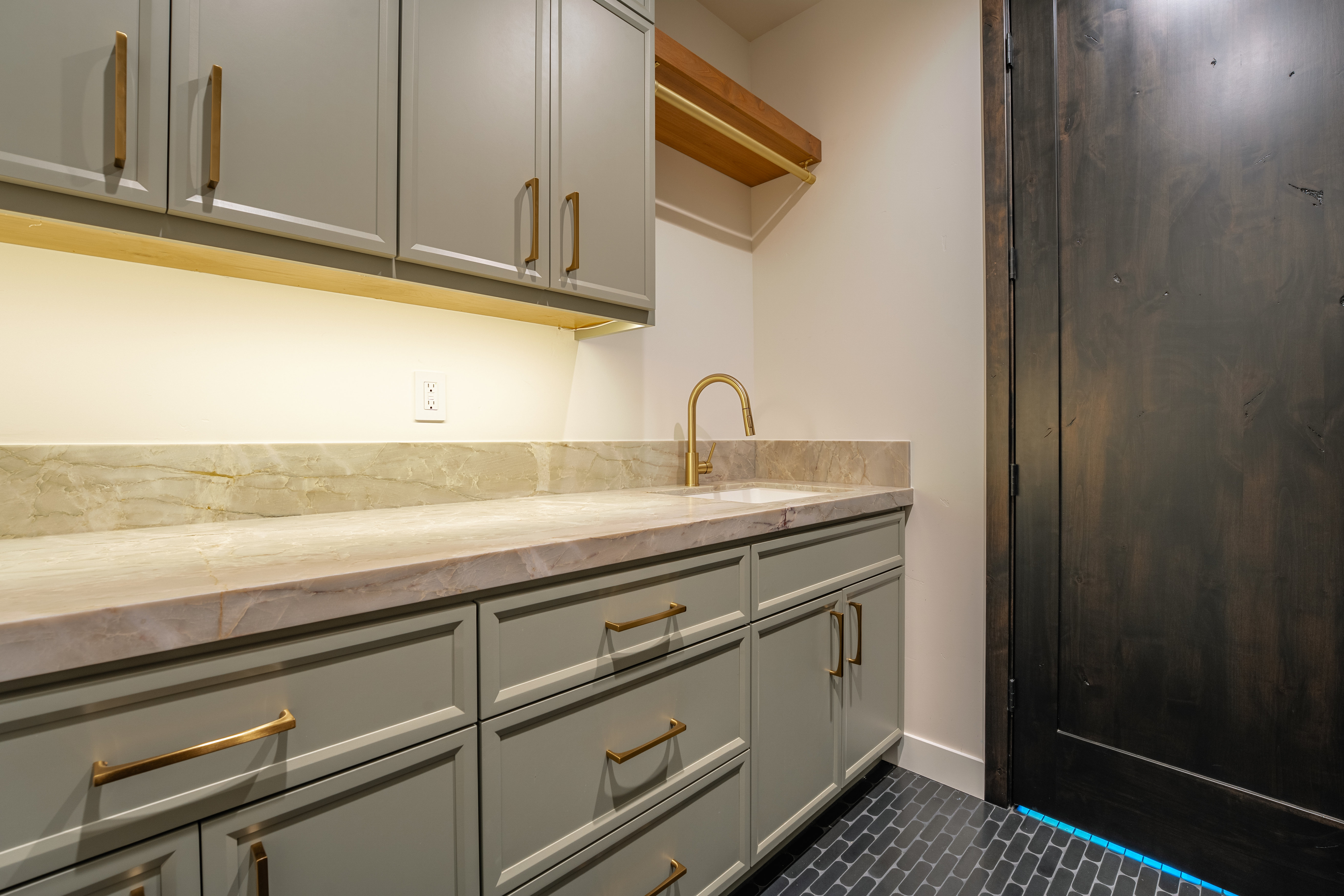 This interior shot showcases a well-appointed laundry room featuring gray cabinetry with gold hardware, a marble countertop, and a gold faucet. The room is illuminated by under-cabinet lighting, and the dark tiled floor adds a touch of sophistication. A dark wooden door is visible on the right, contributing to the room's modern aesthetic.