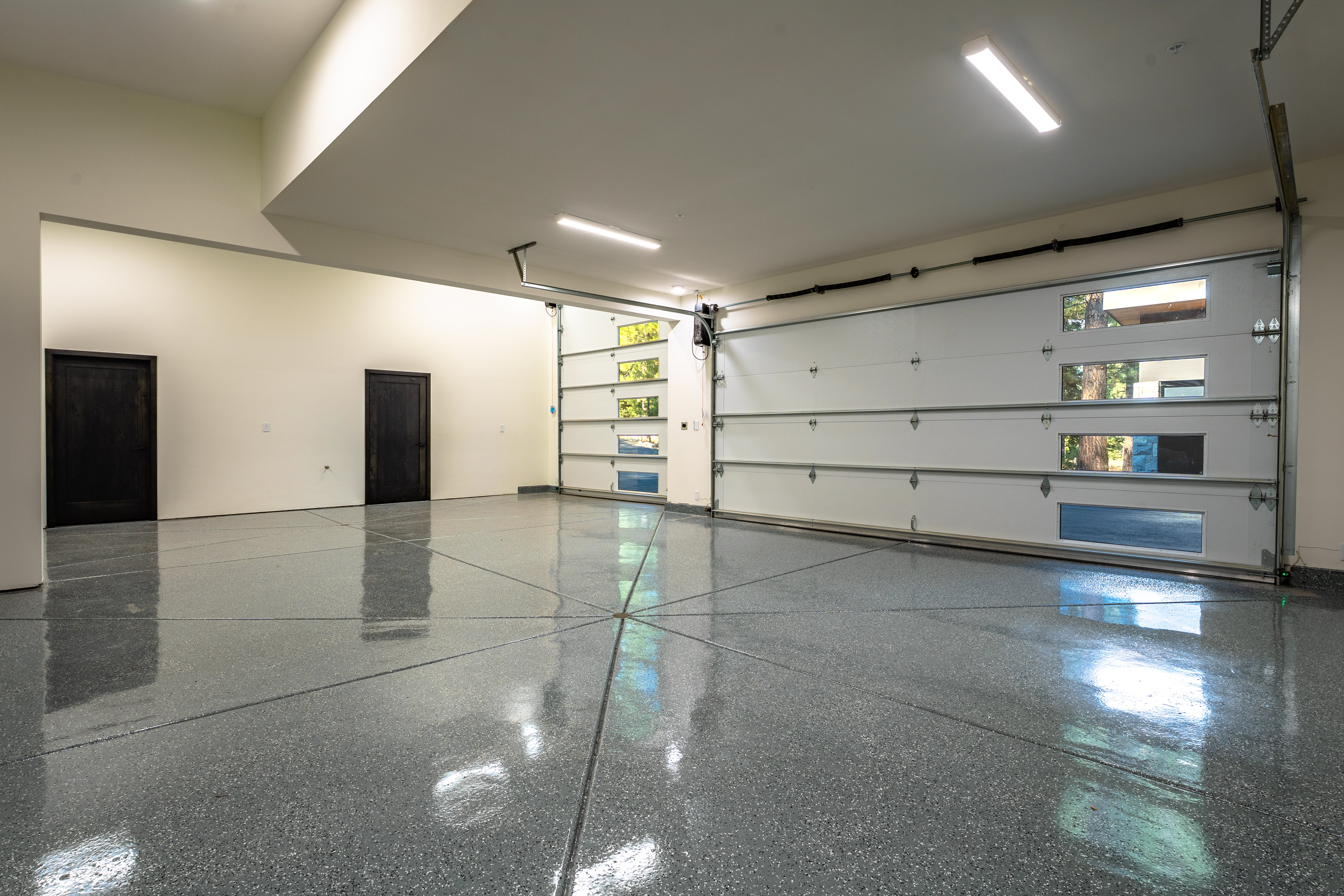 This is a spacious and well-lit garage featuring a modern design with a polished, speckled epoxy floor. The garage door is partially open, revealing a glimpse of the outdoors through its window panels. Two dark-colored doors are visible on the left wall, suggesting access to the main house or additional storage.