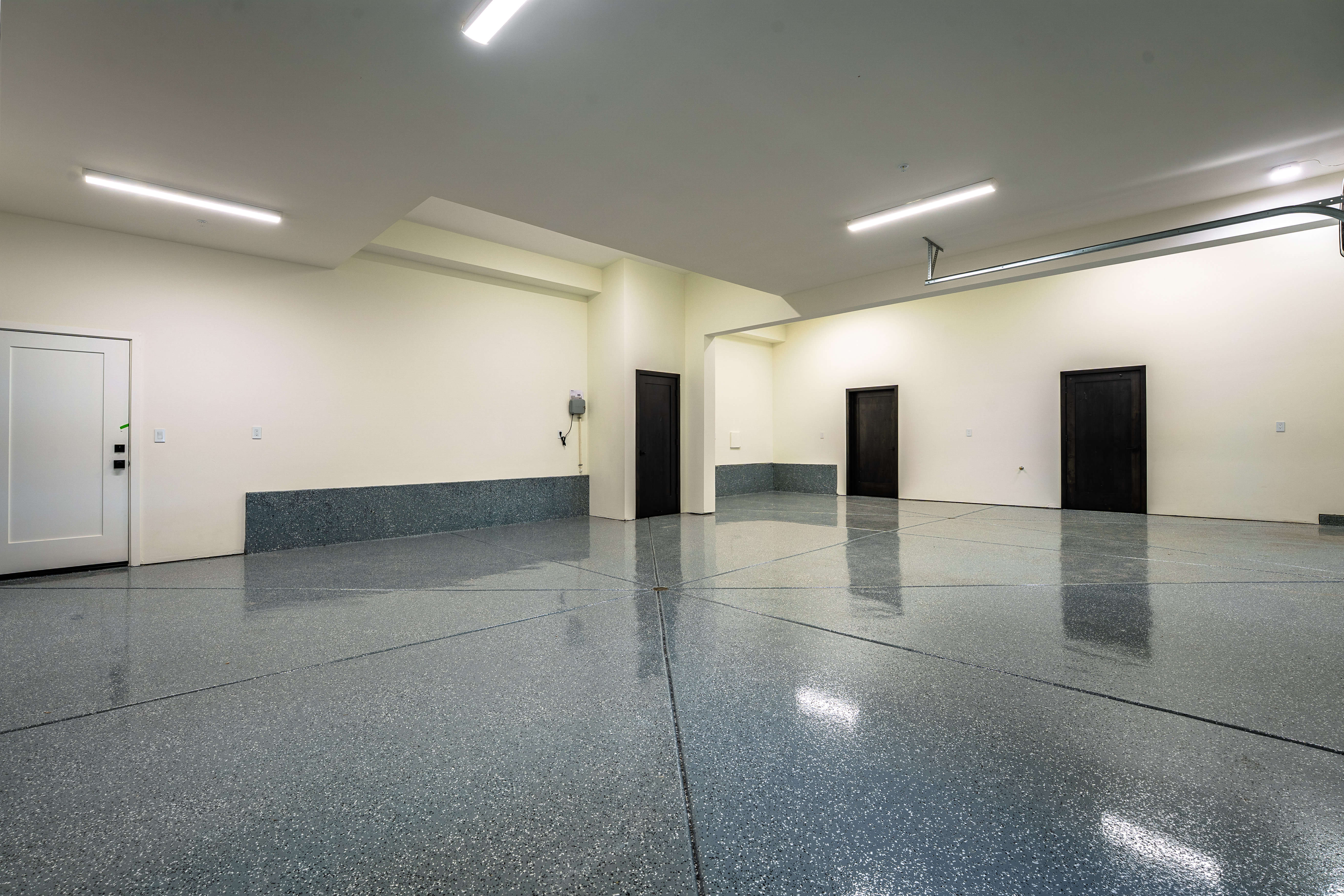 This is a spacious and well-lit garage with a modern aesthetic. The floor has a speckled epoxy finish, and the walls are painted in a neutral tone with a darker accent band. There are three dark-colored doors and one white door, suggesting ample storage or access to other parts of the house. The perspective is a wide shot, showcasing the size and cleanliness of the garage.